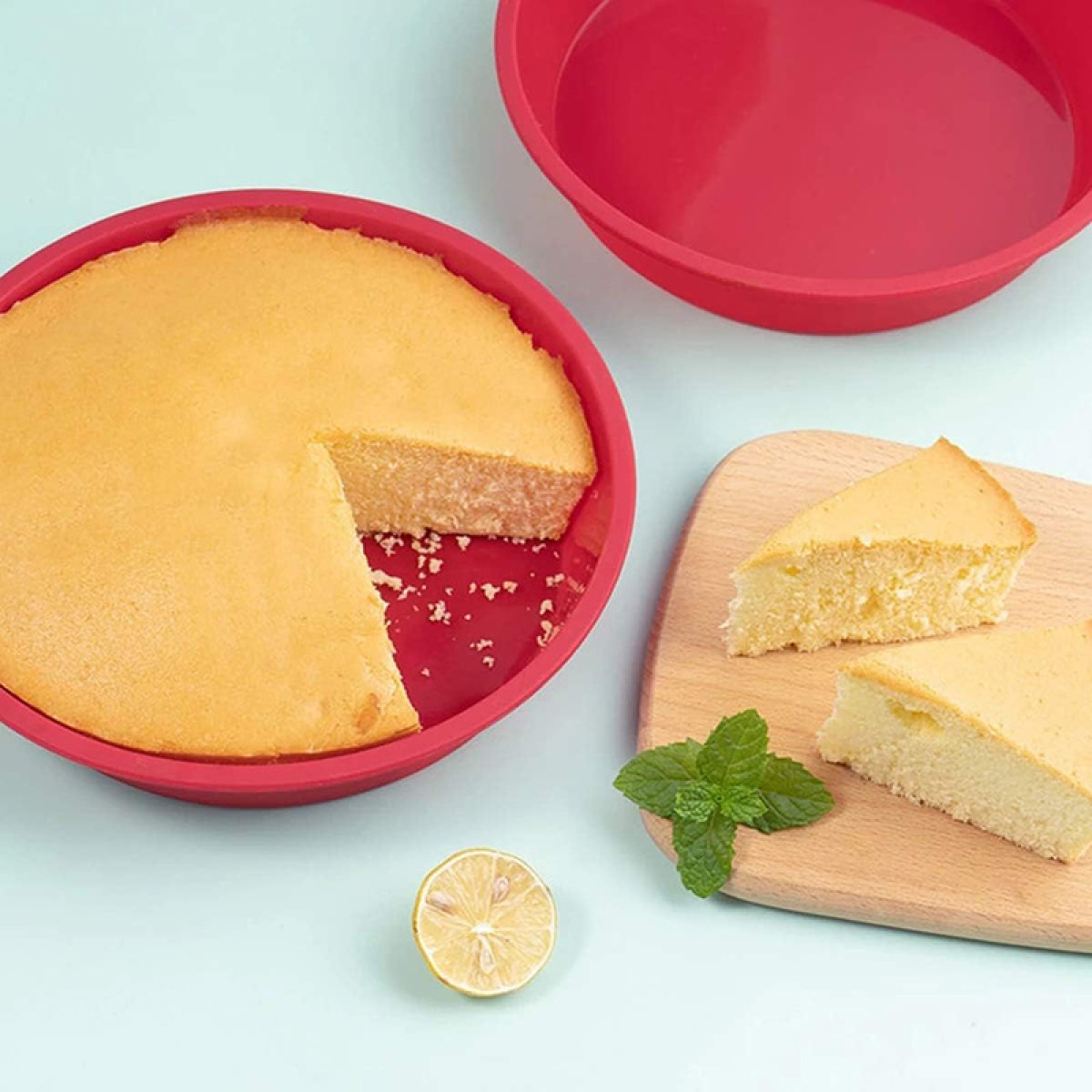 Ein Kuchen mit fehlender Scheibe in einer roten Pfanne, mit Scheiben auf einem Holzbrett, Minze und einer Zitronenscheibe.