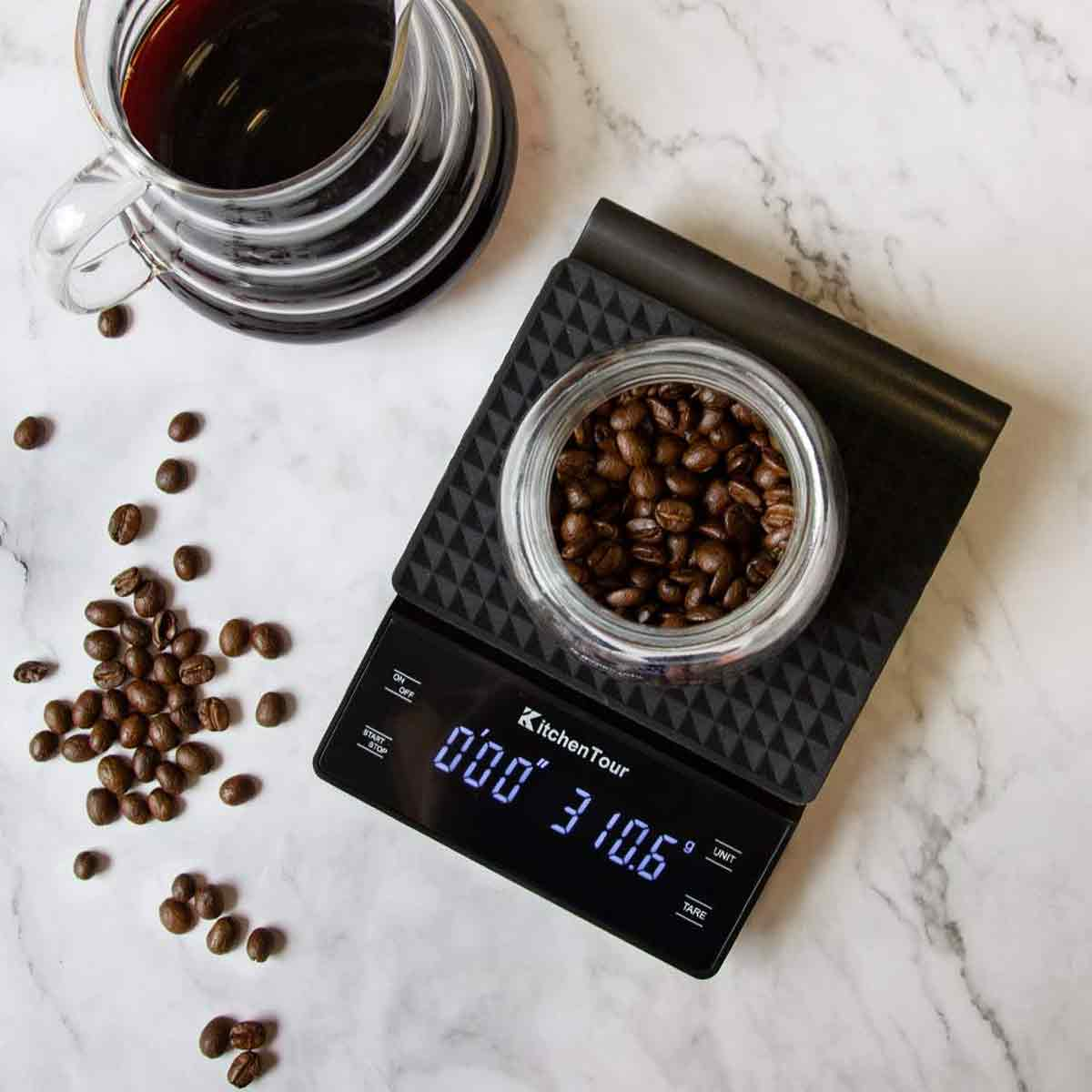 Ein Kaffee-Setup mit Waage, Bohnen und einer Glaskaraffe mit Kaffee auf einer Marmoroberfläche.
