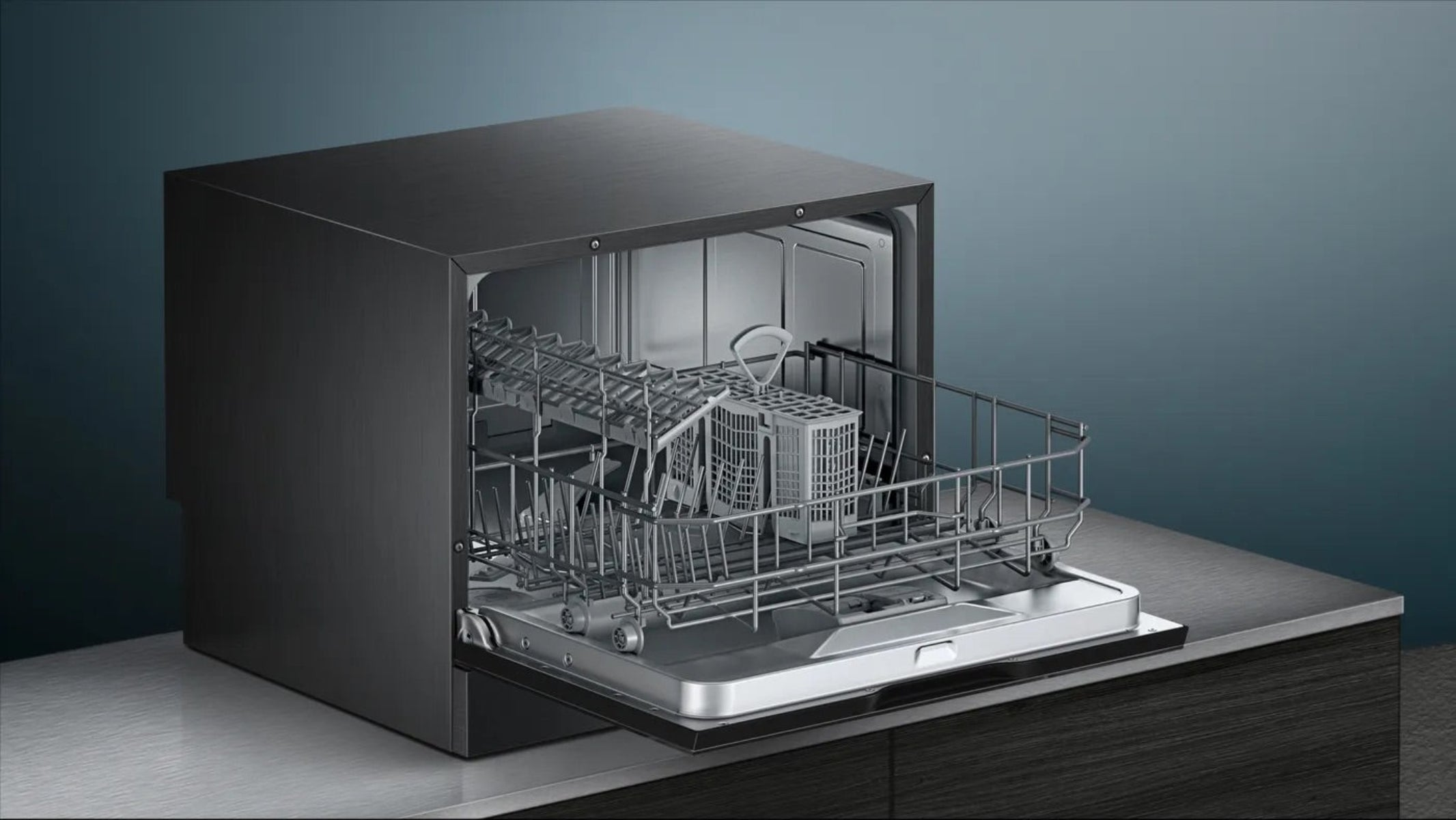Ciemnoszara zmywarka z otwartymi drzwiami, odsłaniająca wewnętrzne kosze i koszyk. Stoi na ciemnym blacie.