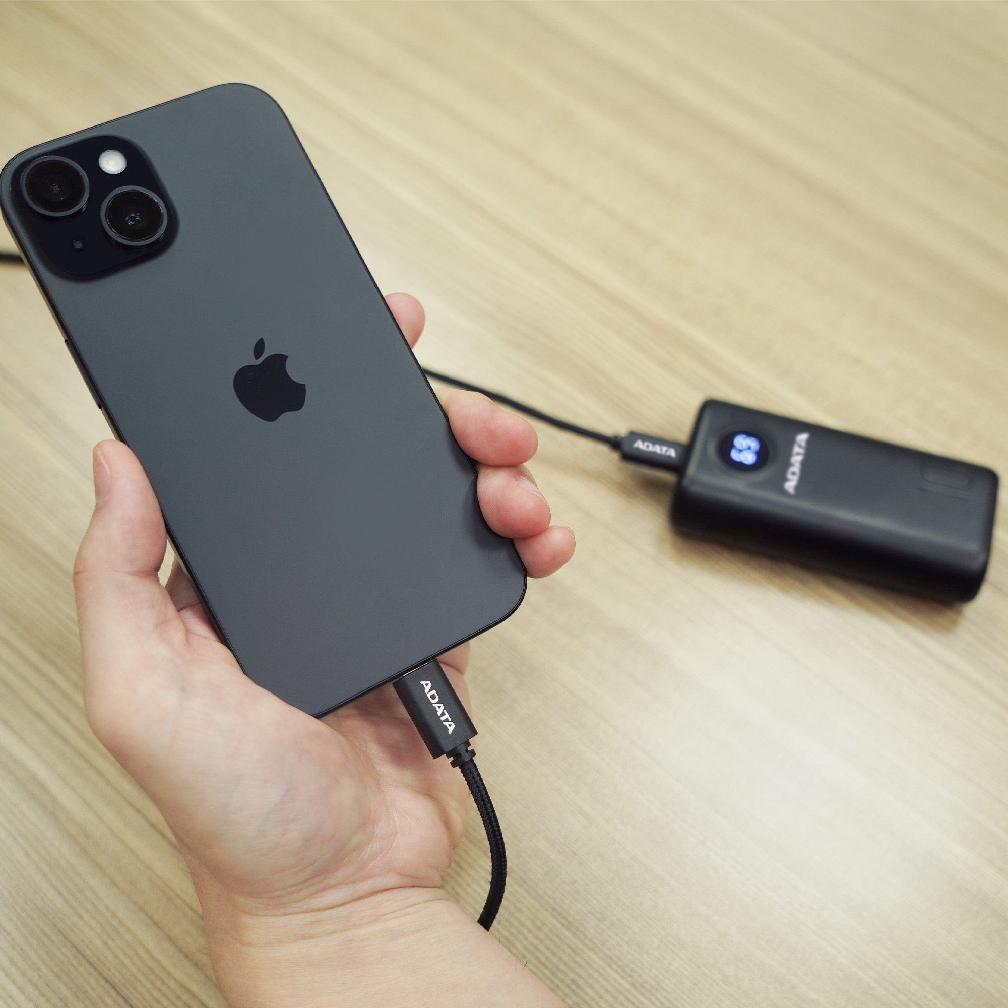 Eine Hand, die ein schwarzes iPhone hält, das mit einer Powerbank verbunden ist. Die Powerbank zeigt die restliche Batterie.