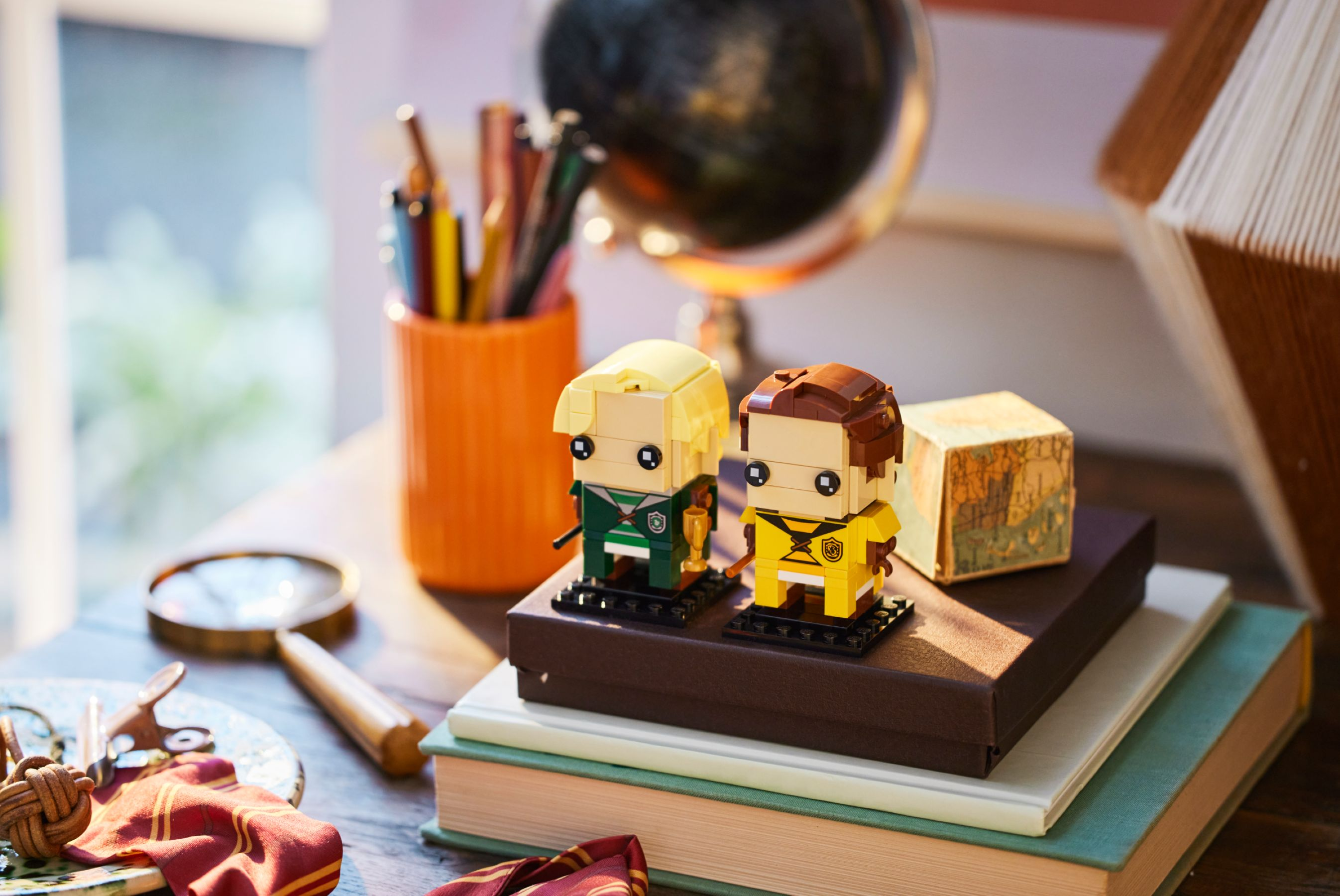 Zwei Lego-Figuren auf Büchern. Eine ist grün und gold, die andere gelb und braun. Daneben sind ein Globus und Bleistifte.