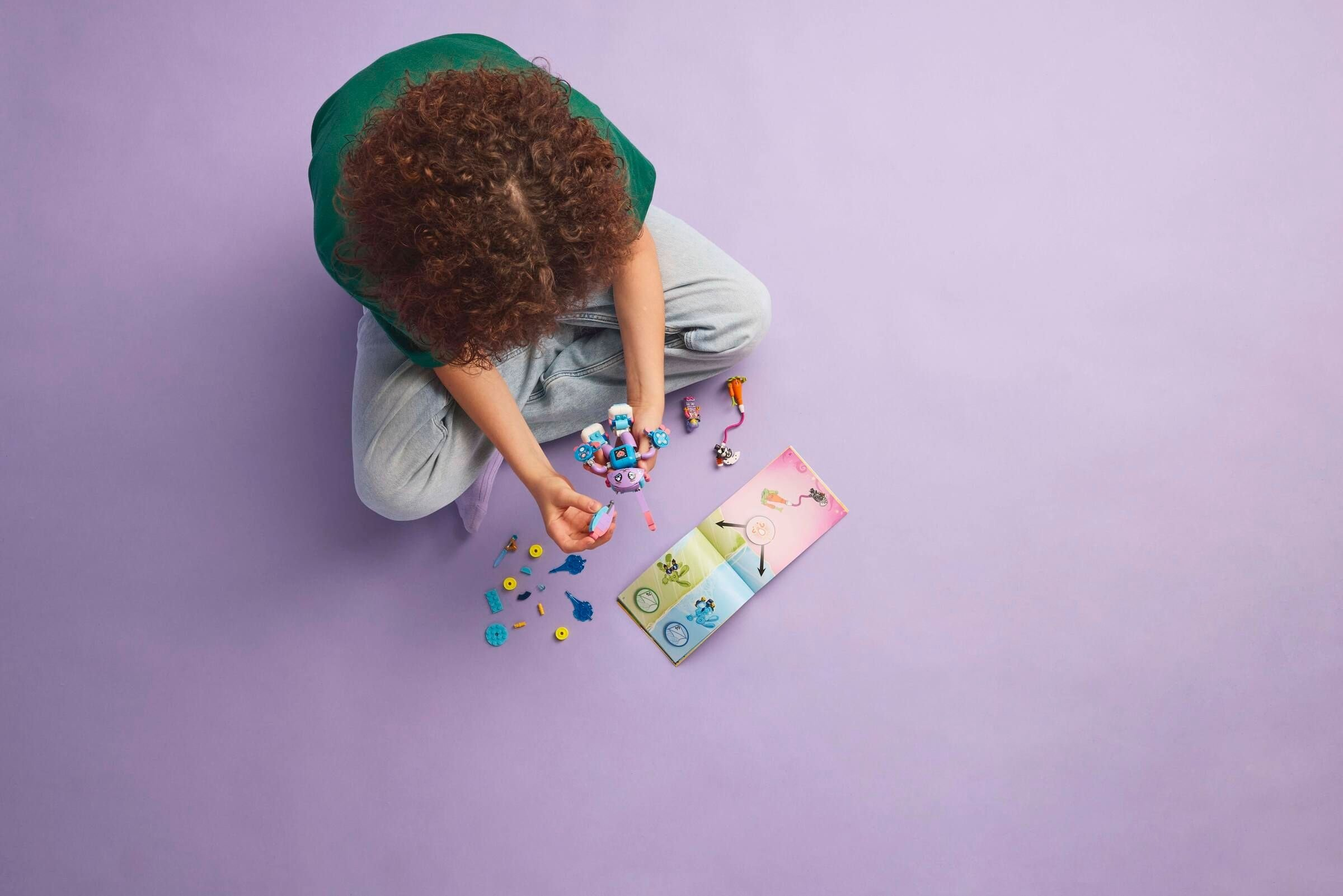 Eine Person spielt mit LEGOs. Sie sind zentriert mit einem lila Hintergrund. Rechts befindet sich ein Heft.