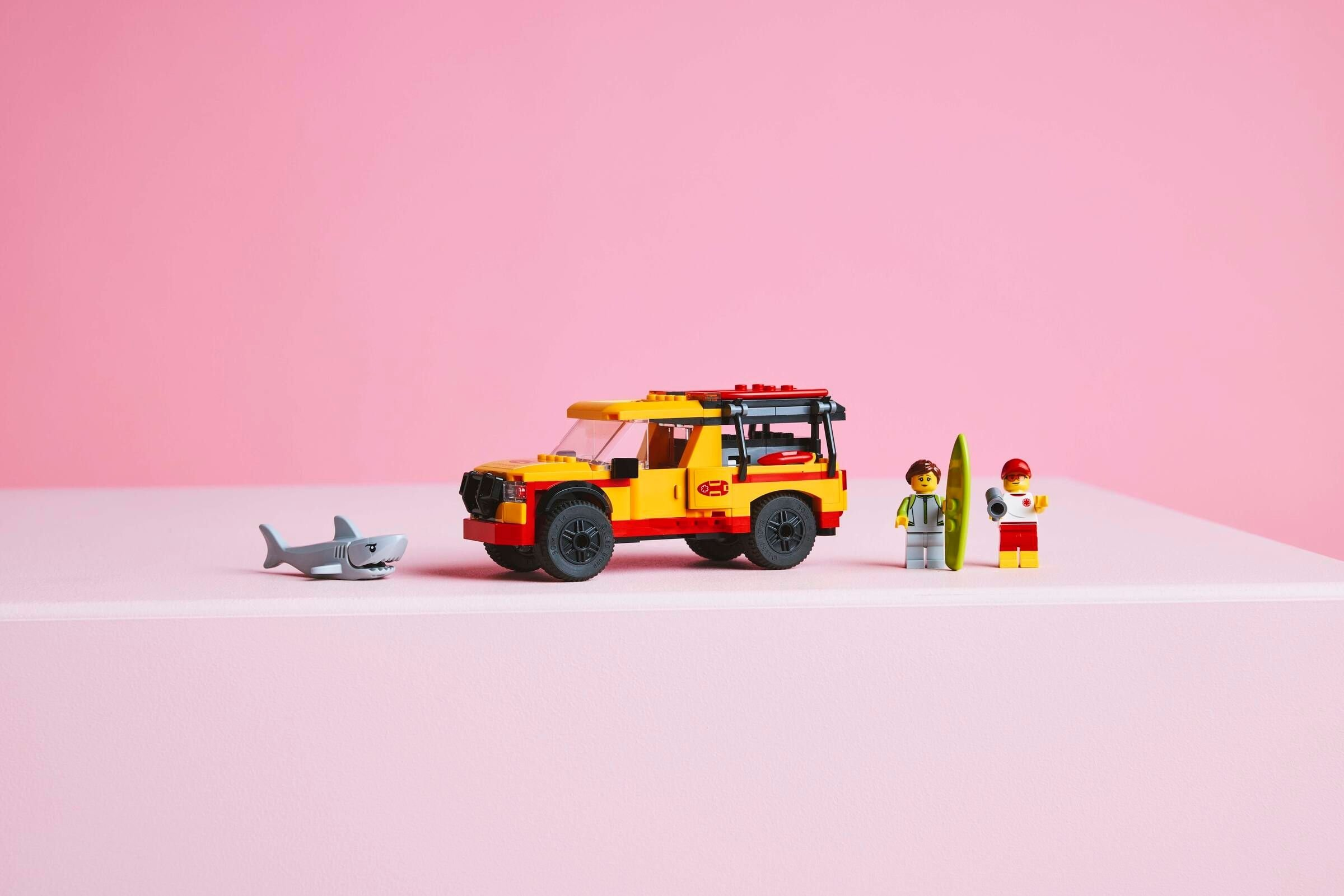 Ein gelber Lego-Rettungswagen, Hai, zwei Figuren und Surfbrett auf rosa Hintergrund.