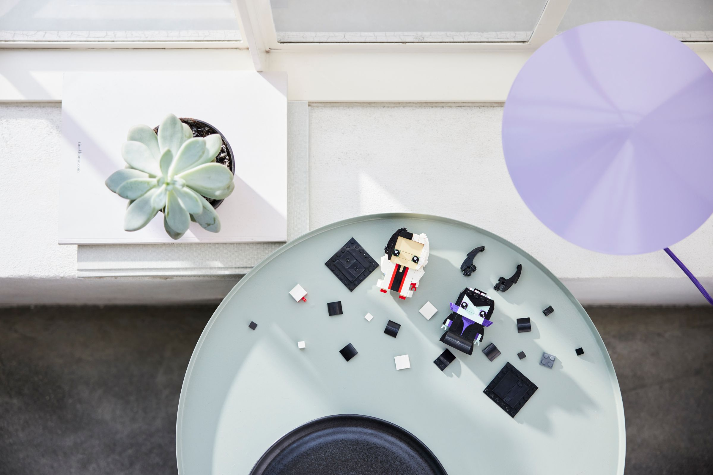Draufsicht auf einen Tisch mit Lego-Figuren und einem Ballon. Im Hintergrund sind eine Pflanze und ein Fenster zu sehen.