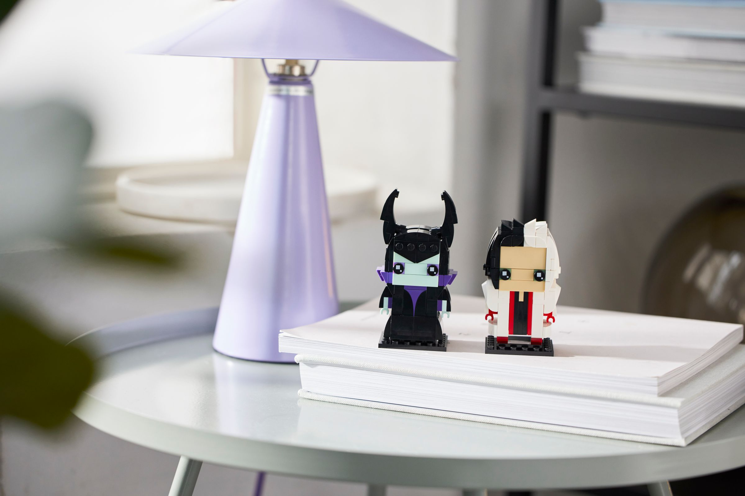 Zwei Lego-Figuren von Disney-Bösewichten, Maleficent und Cruella, sitzen auf einem Buch. Auf dem Tisch steht eine lila Lampe.