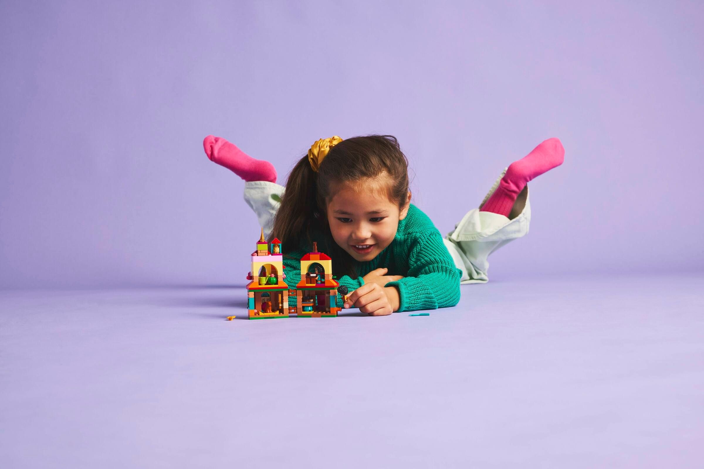 Ein Mädchen liegt auf Lila und spielt mit einem Lego-Schloss. Sie trägt einen grünen Pullover, rosa Socken. Das Schloss ist bunt.