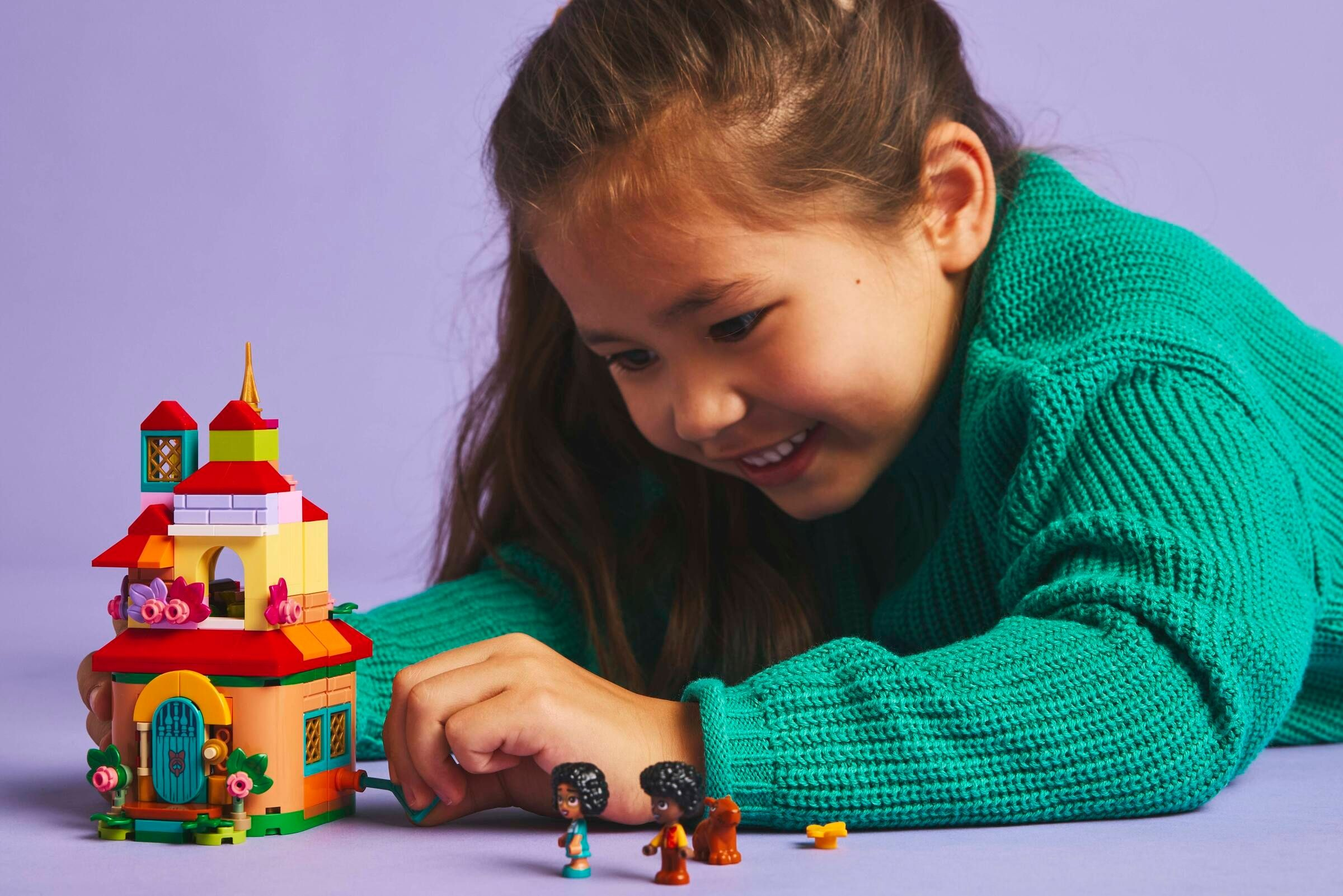 Ein Mädchen lächelt ein farbenfrohes Lego-Haus an. Zwei kleine Figuren und ein braunes Tier sind platziert.