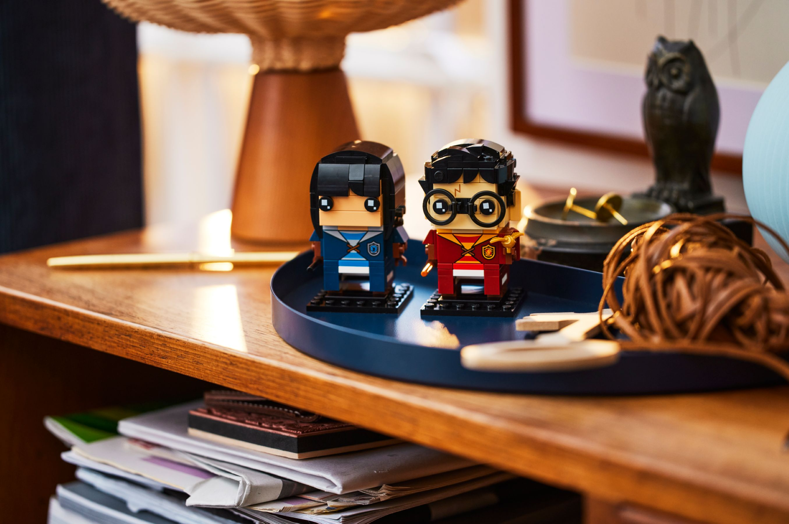 Zwei Lego-Figuren von Harry-Potter-Charakteren auf einem blauen Tablett auf einem Holztisch. Eine dekorative Eule ist im Hintergrund.