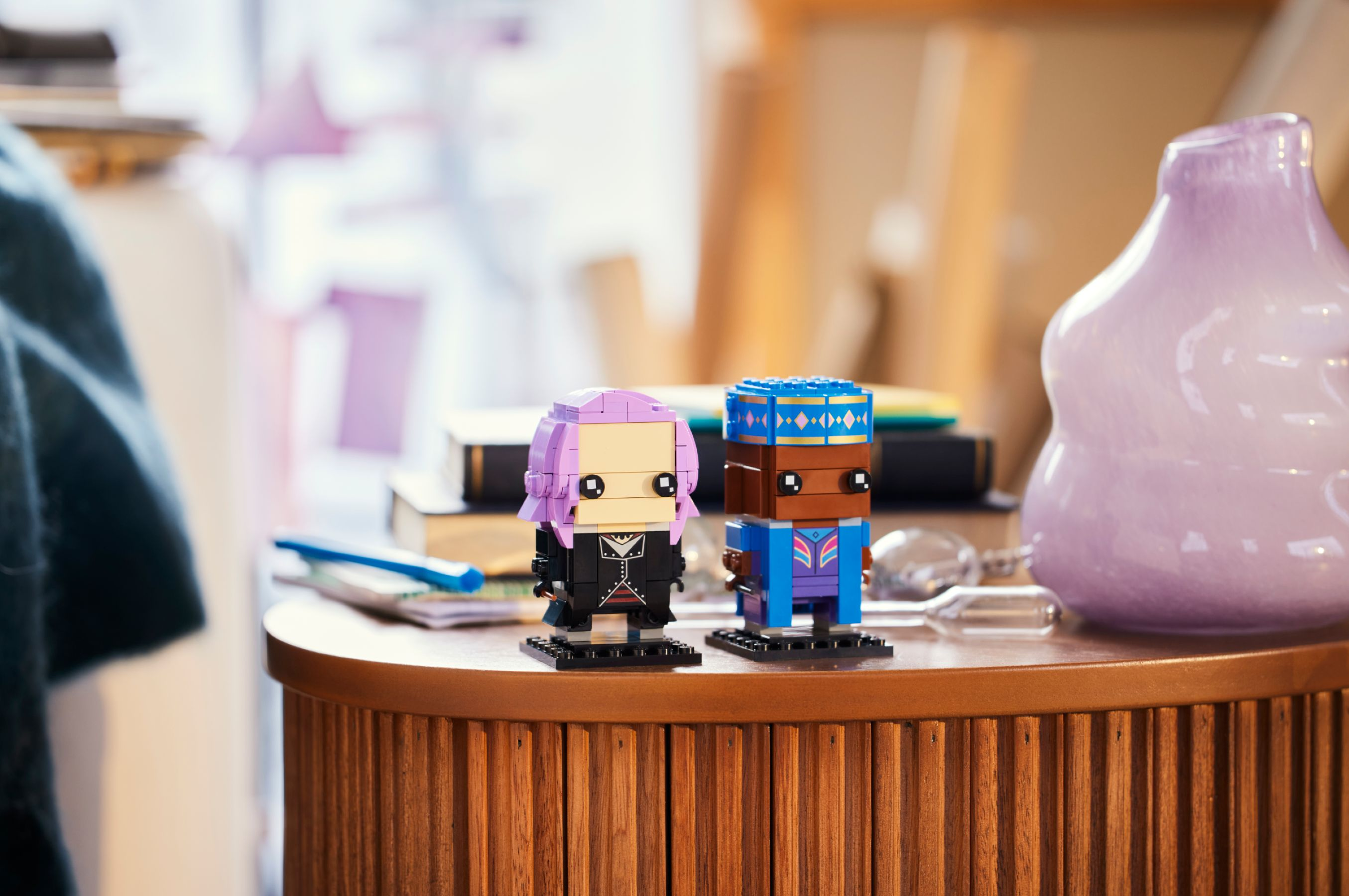 Zwei Lego-Figuren mit lila und blauen Haaren auf einem Holztisch. Im Hintergrund steht eine lila Vase.