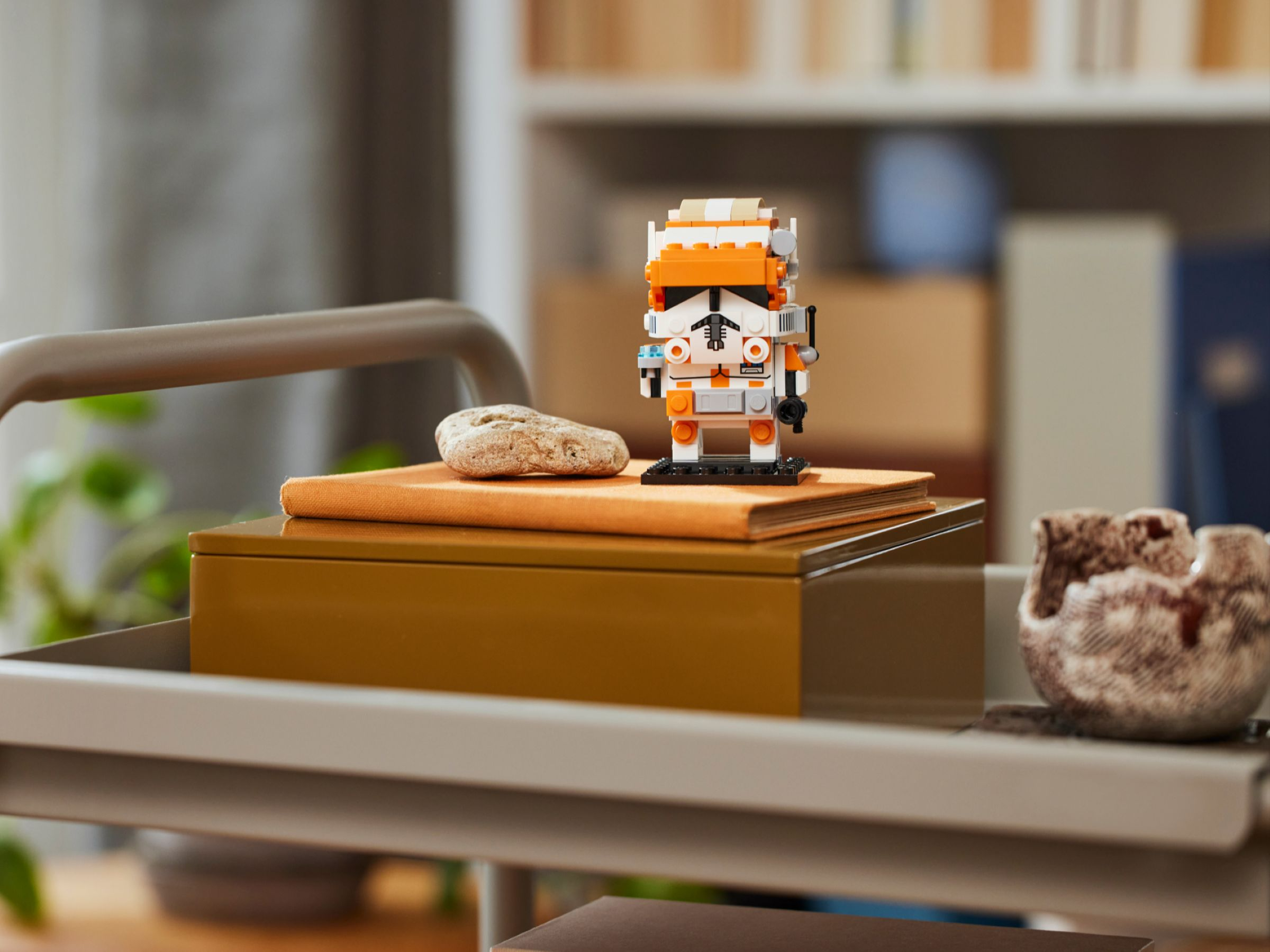 Eine Lego-Figur eines Klonkriegers auf einem Tisch, mit einem Stein und einer dekorativen Schüssel. Bücherregale sind im Hintergrund.