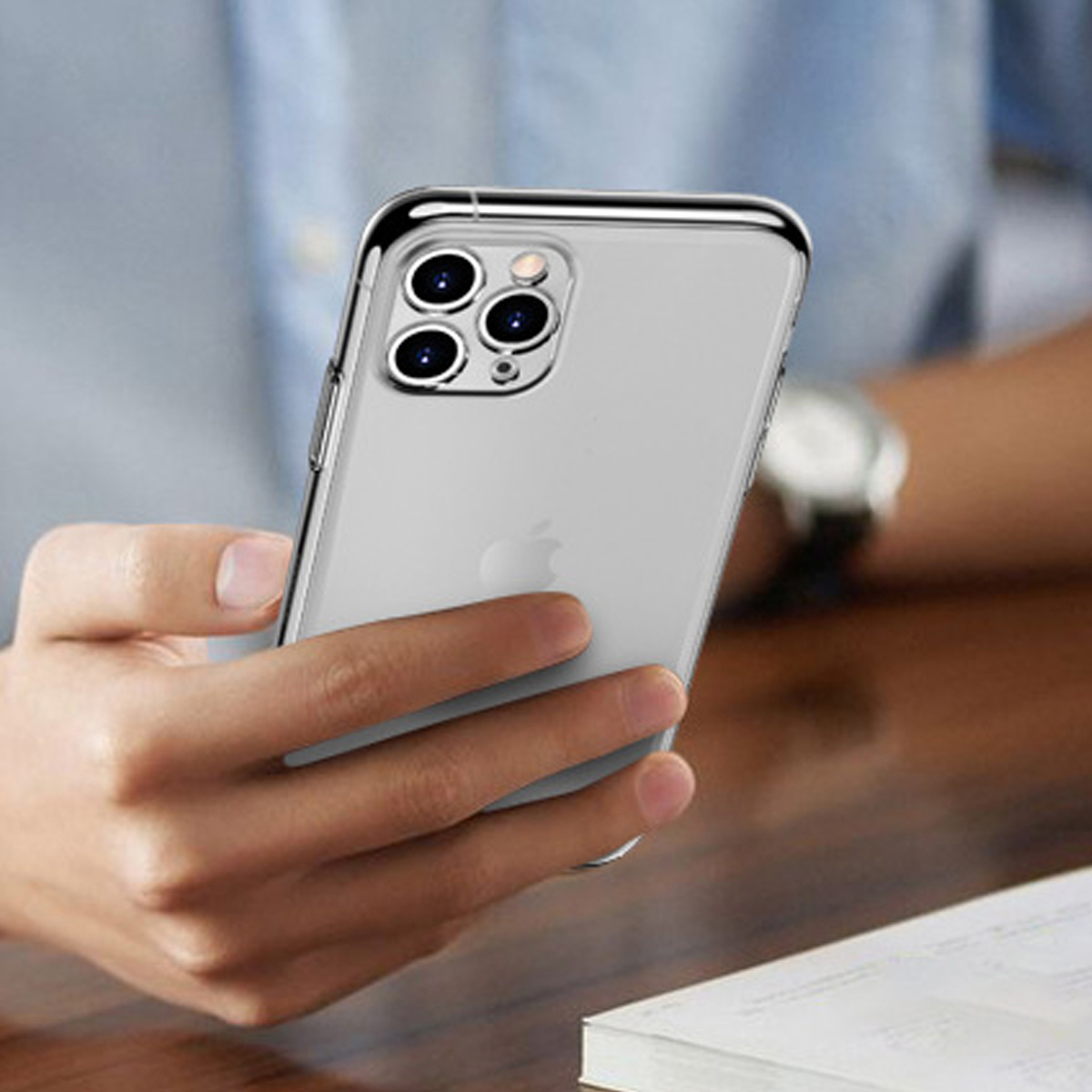Eine Person hält ein iPhone in einer transparenten Hülle. Das Telefon ist silberfarben mit einem weißen Apple-Logo. Der Hintergrund ist unscharf.