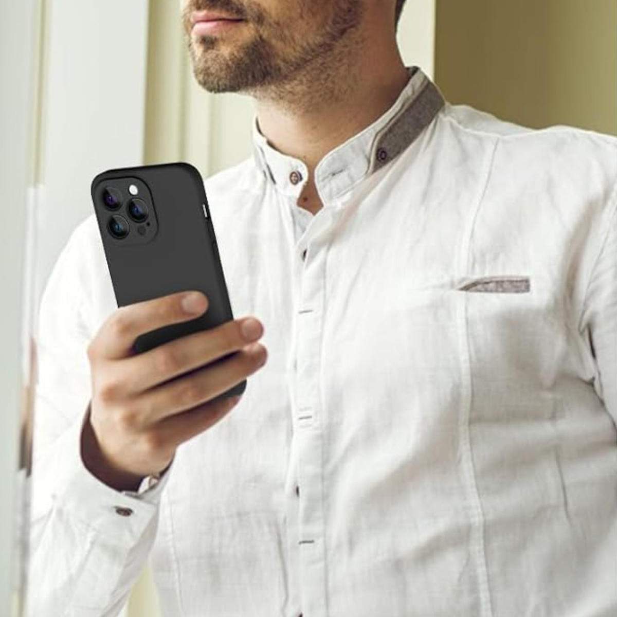 Ein Mann in weißem Hemd hält ein schwarzes Telefon. Das Telefon hat drei Kameralinsen. Der Mann hat einen Bart und steht an einem Fenster.