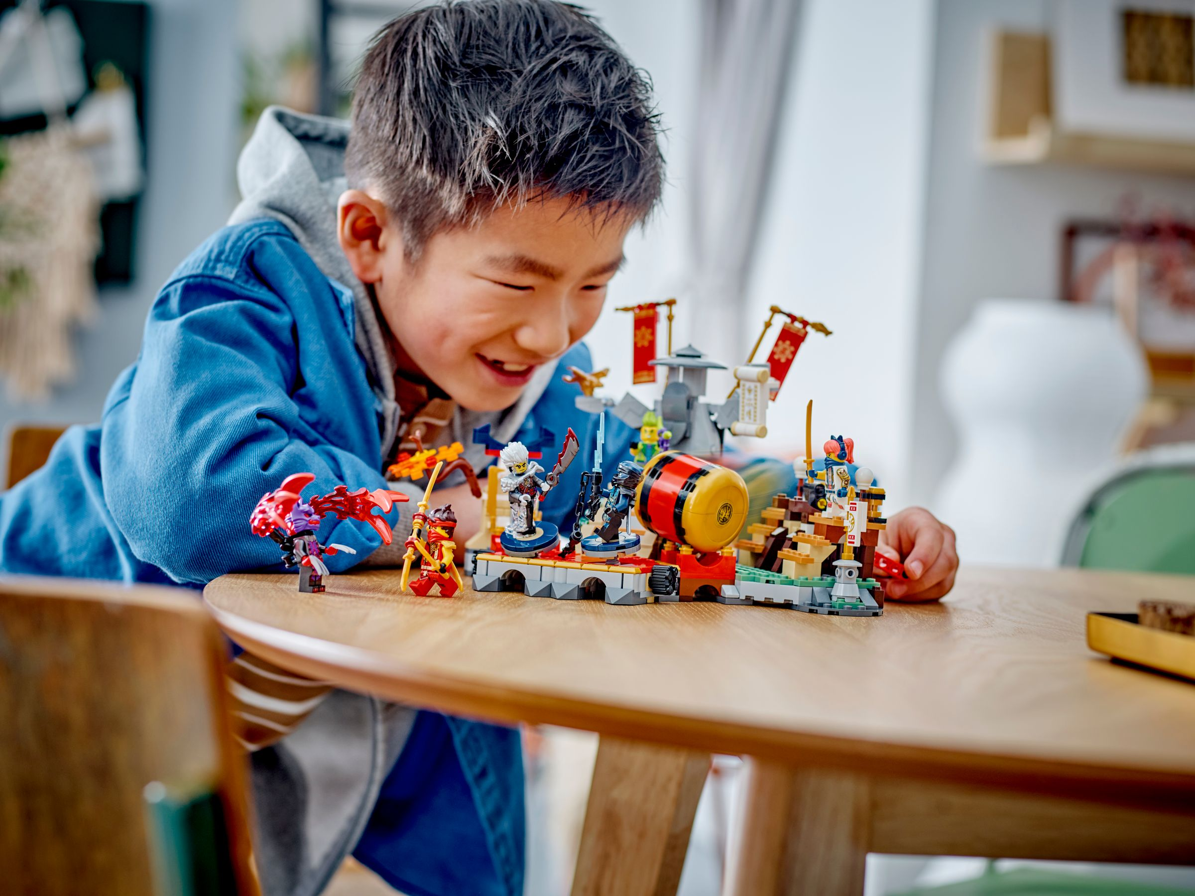 Ein Junge lächelt über ein Lego Ninjago Set. Das Set zeigt Figuren, Gebäude und eine große Trommel auf einem Holztisch.