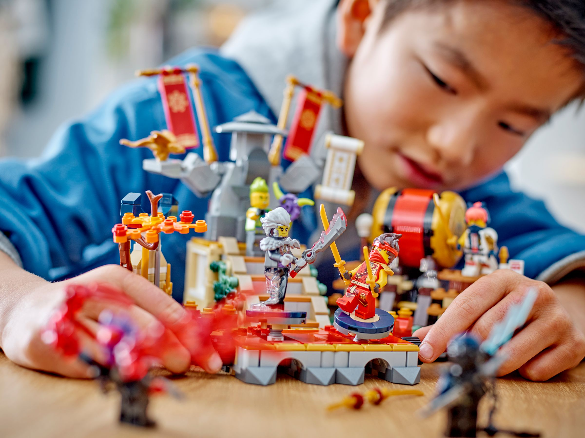 Ein Kind spielt mit Lego Ninjago. Mehrere Figuren und Strukturen befinden sich auf einem Holztisch. Das Gesicht des Kindes ist sichtbar.