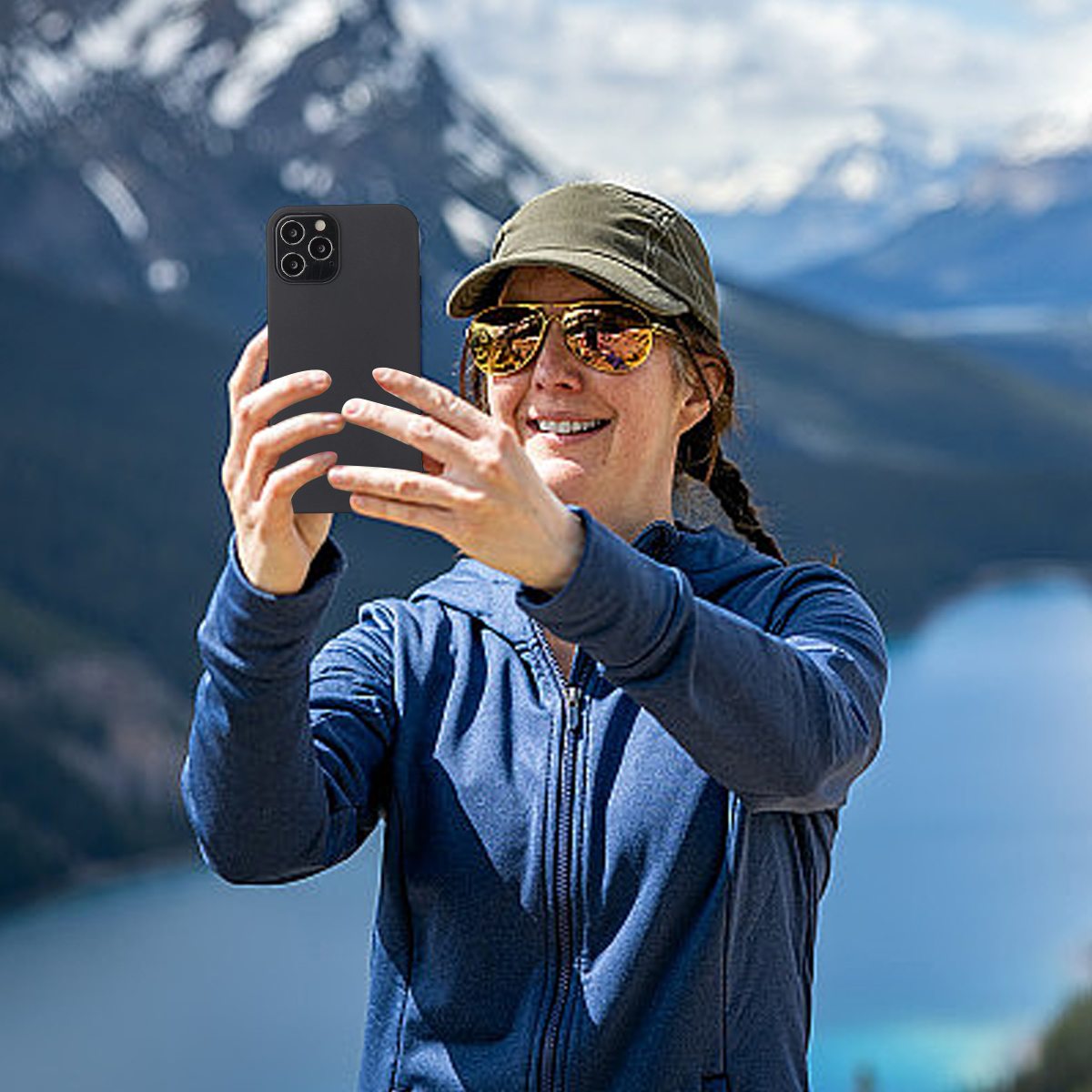 Eine Frau macht ein Selfie mit einem Telefon, trägt einen Hut und eine Sonnenbrille, mit einem See und Bergen im Hintergrund.
