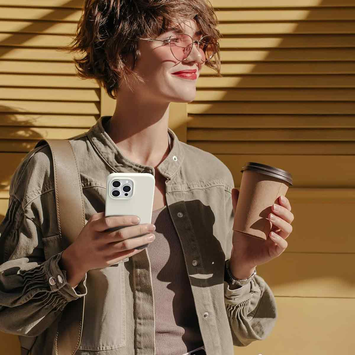 Frau mit Telefon und Kaffee, Brille und Jacke. Gelber Fensterladen im Hintergrund.