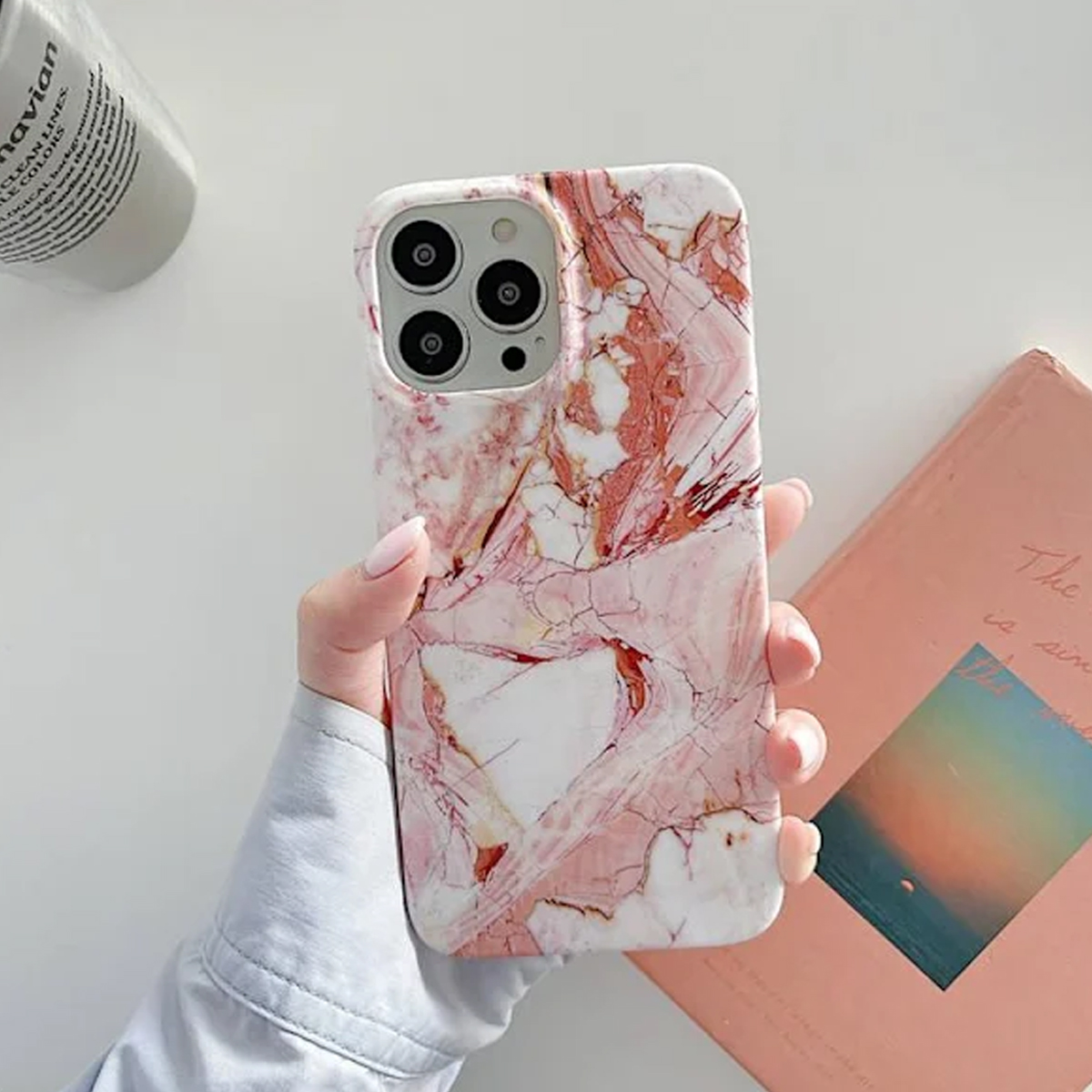 Eine Hand, die eine Marmor-Handyhülle hält, mit einem rosa Buch und einem weißen Objekt im Hintergrund.