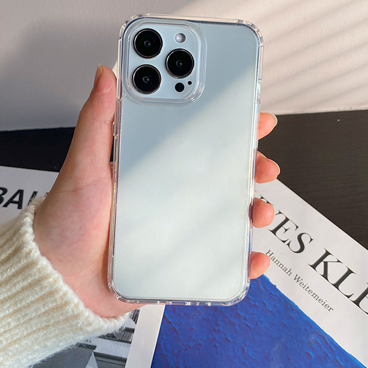 Eine Hand hält ein Telefon mit einer durchsichtigen Hülle. Das Telefon hat drei Kameralinsen. Im Hintergrund ein Buch mit Text.