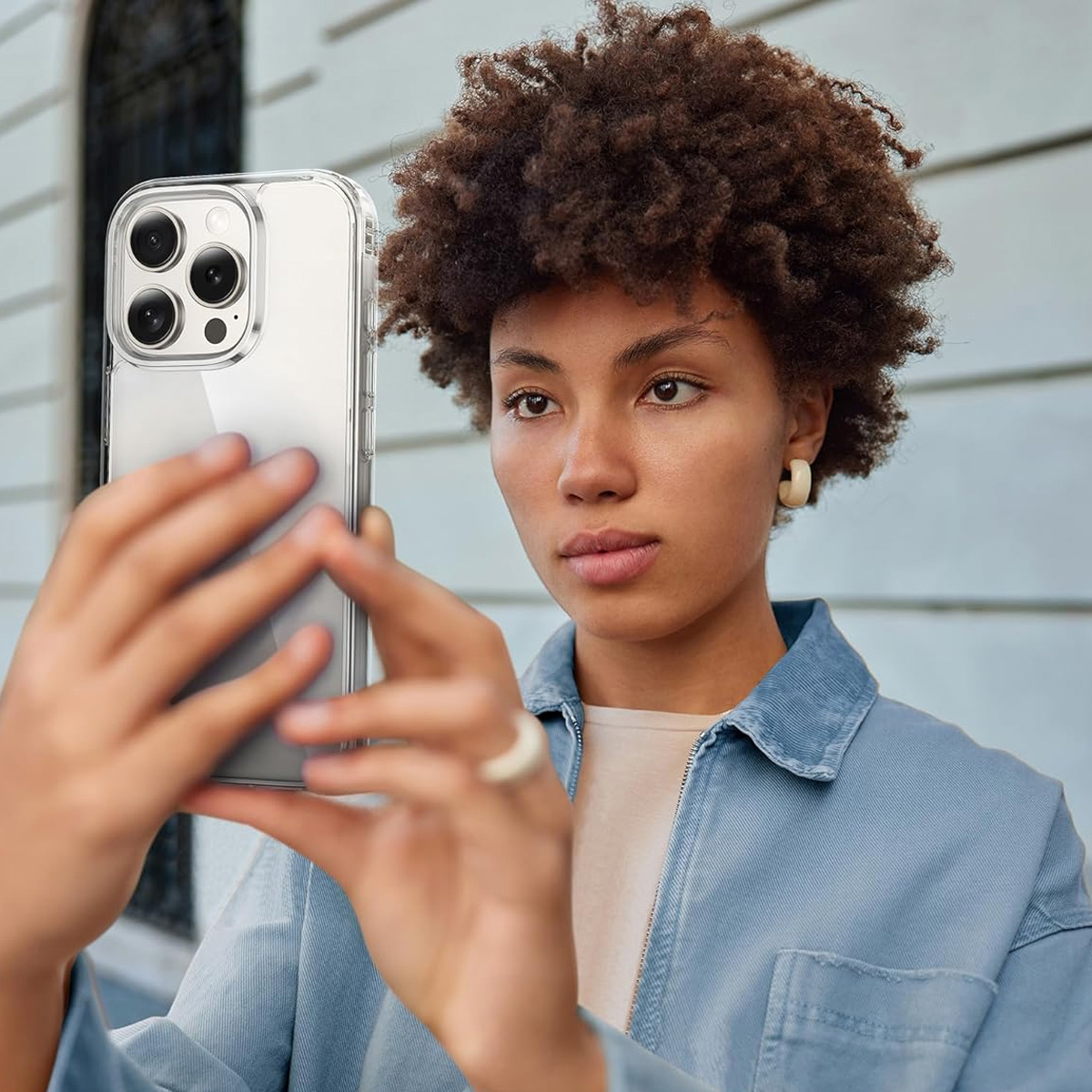 Eine Frau macht ein Selfie mit einem Telefon. Sie hat lockiges Haar, eine Jeansjacke und einen weißen Ring. Hintergrund ist ein Gebäude.