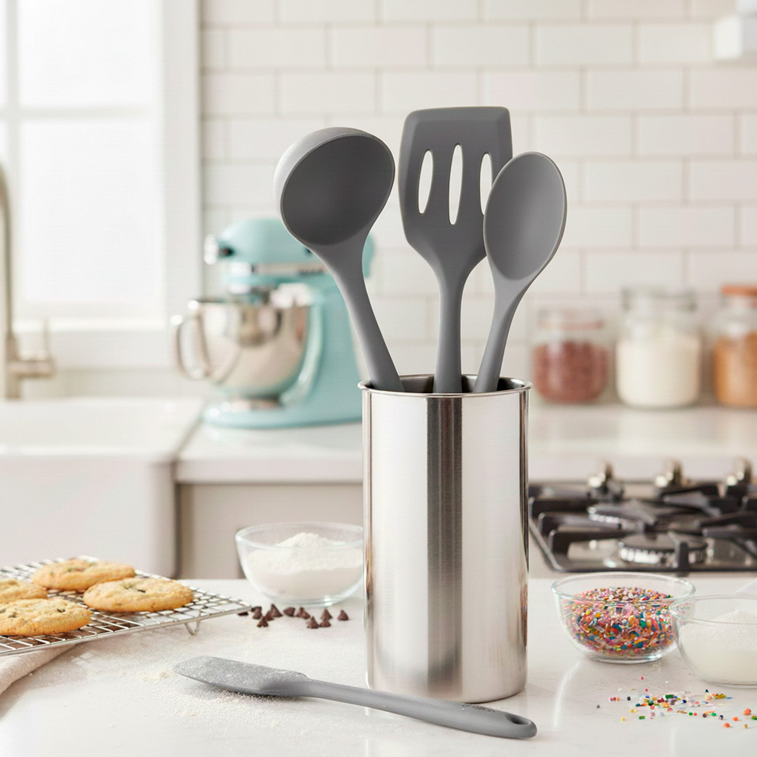 Escena de cocina con utensilios en un soporte de metal, galletas, cuencos con ingredientes y una batidora en el fondo.