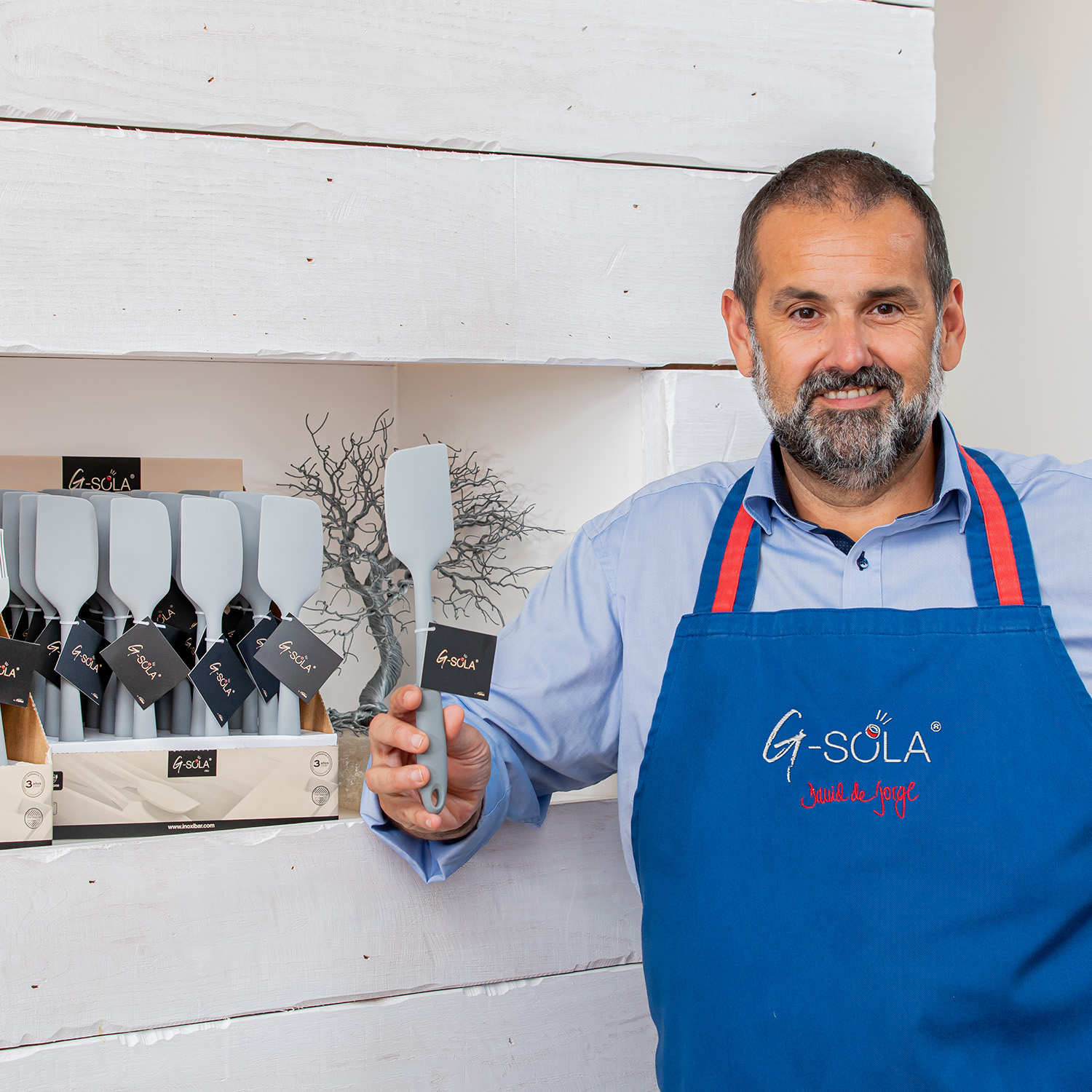 Un hombre con un delantal azul con el logo G-Sola sostiene una espátula gris. Detrás de él hay estantes con espátulas. Pared de madera blanca.