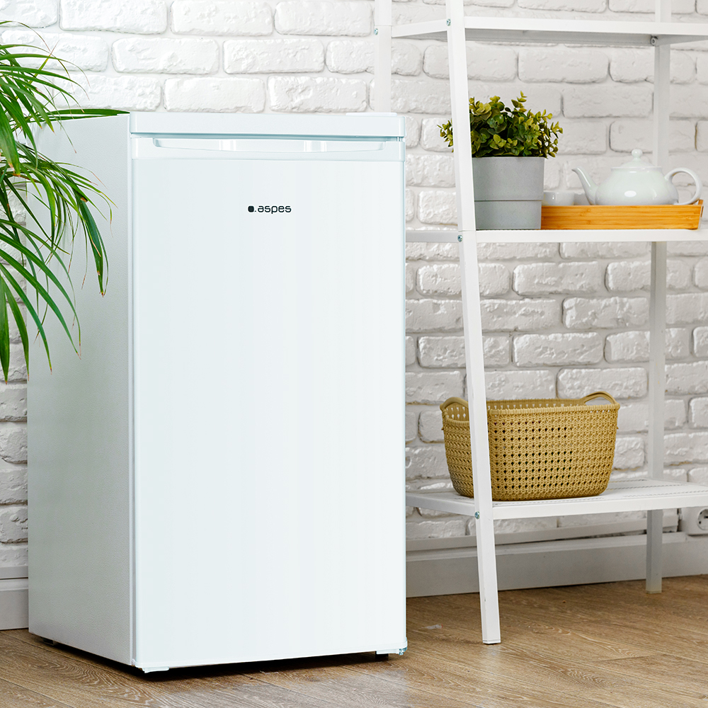 Un refrigerador blanco se encuentra junto a una estantería con una cesta y plantas. Una pared de ladrillo sirve de fondo.