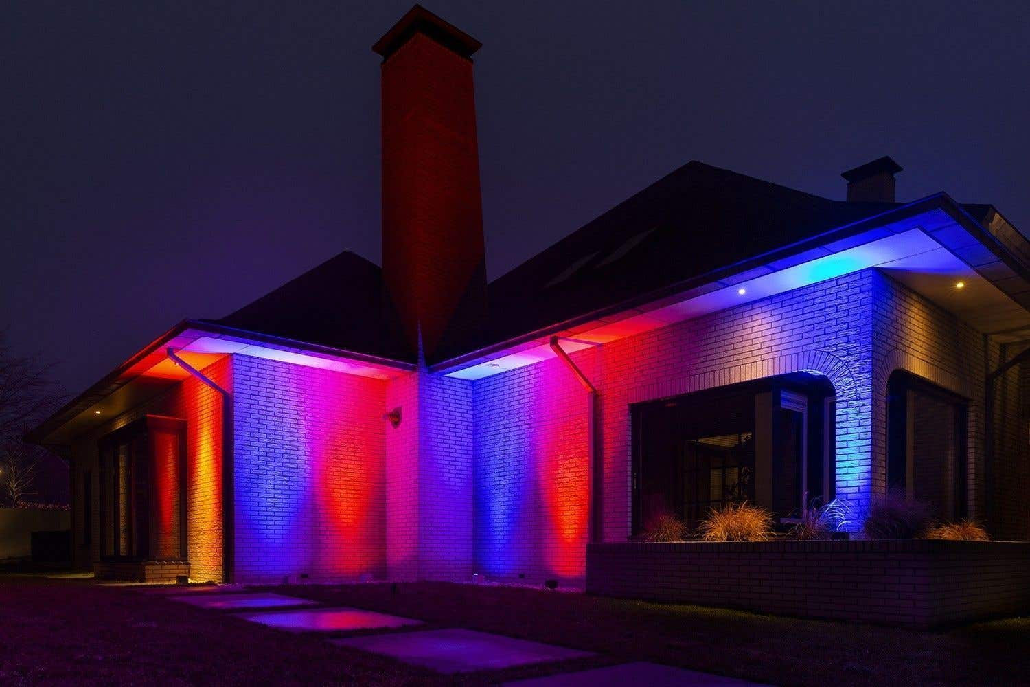 La fachada de una casa está iluminada con luces rojas, azules y amarillas por la noche. Una chimenea roja se alza sobre el tejado. El cielo está oscuro.