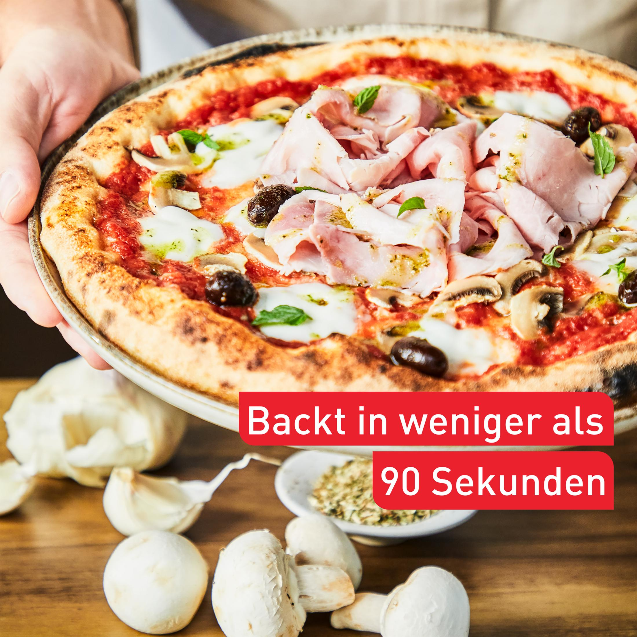 Eine Pizza mit Schinken, Oliven und Pilzen wird gehalten. Knoblauch und Pilze liegen auf einem Holztisch.
