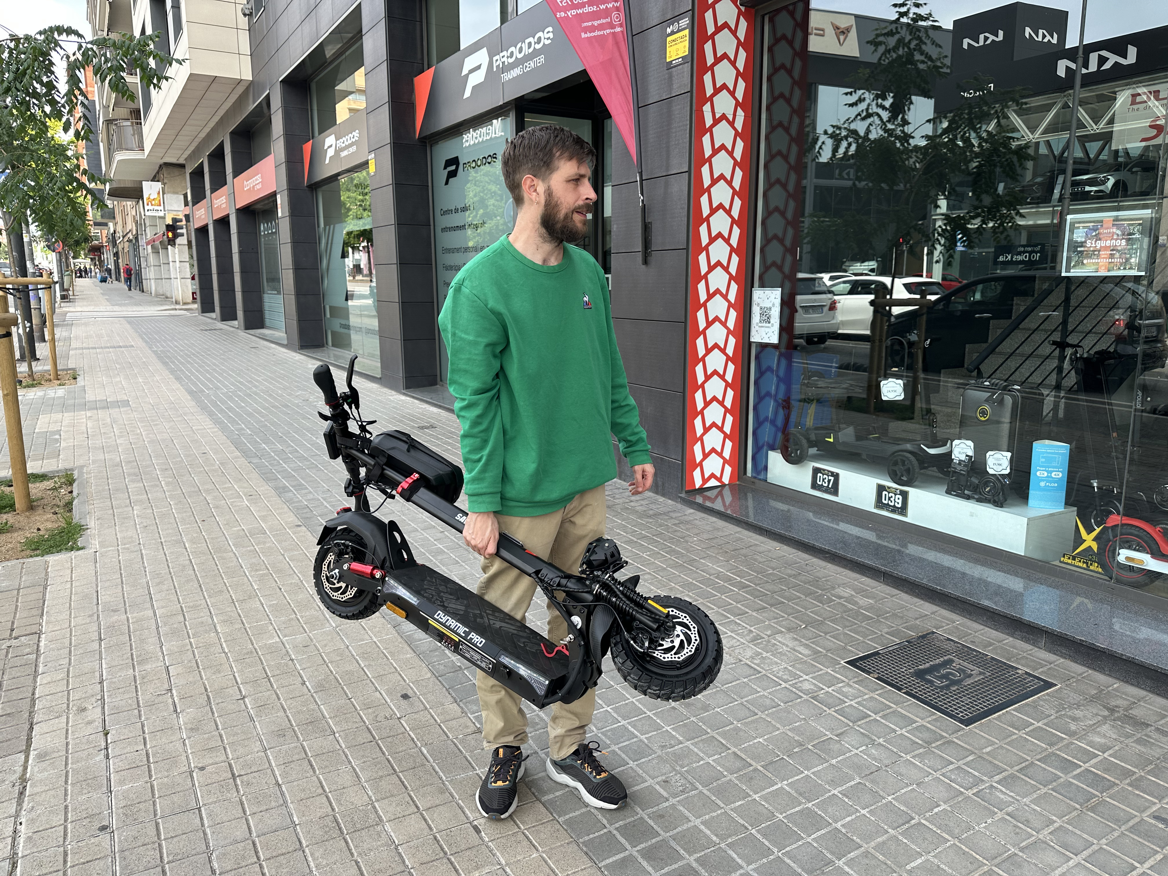 Hombre sosteniendo un scooter negro en una acera. Edificios y tiendas al fondo.
