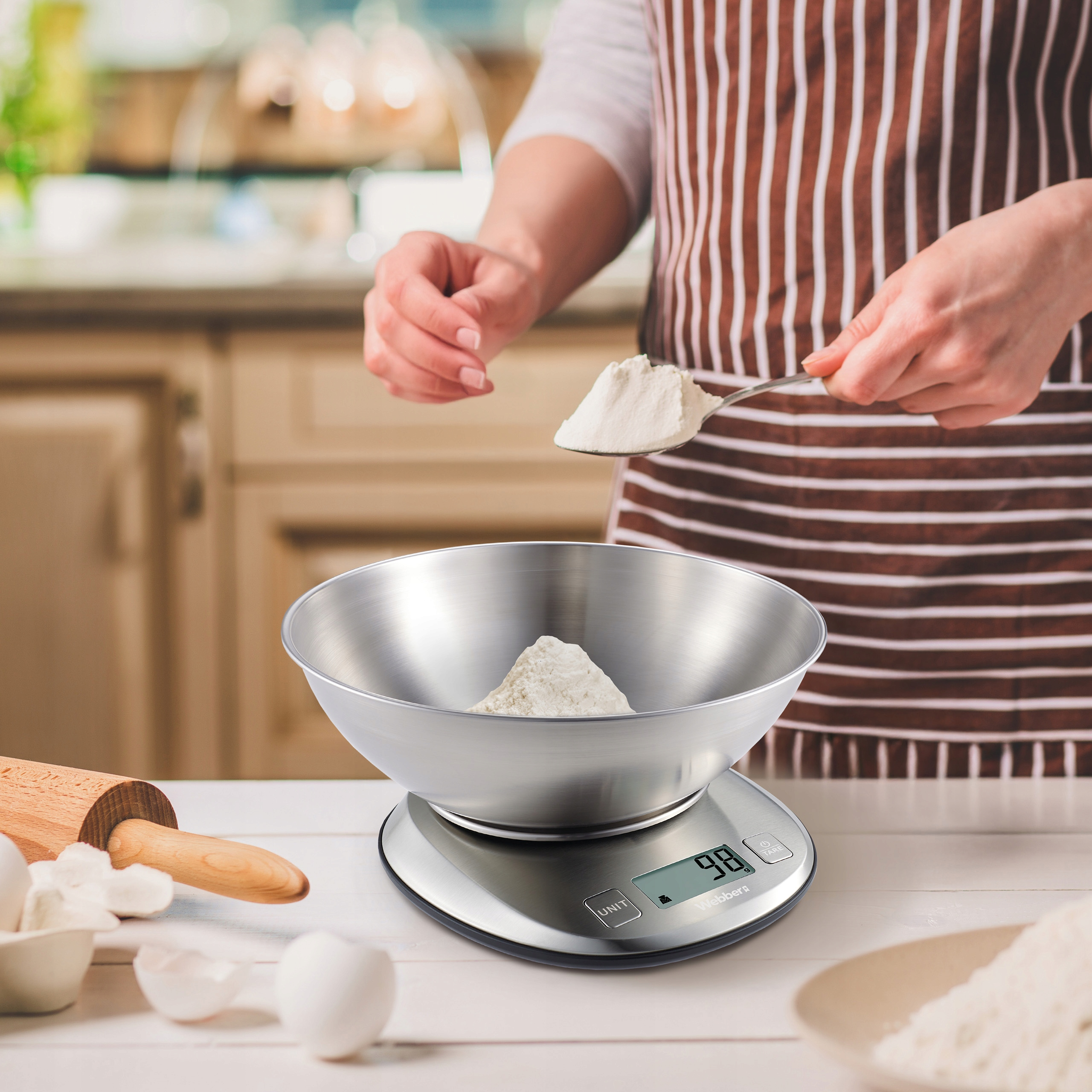 Osoba w fartuchu mierzy mąkę łyżką na wadze kuchennej. Widoczne są inne składniki.