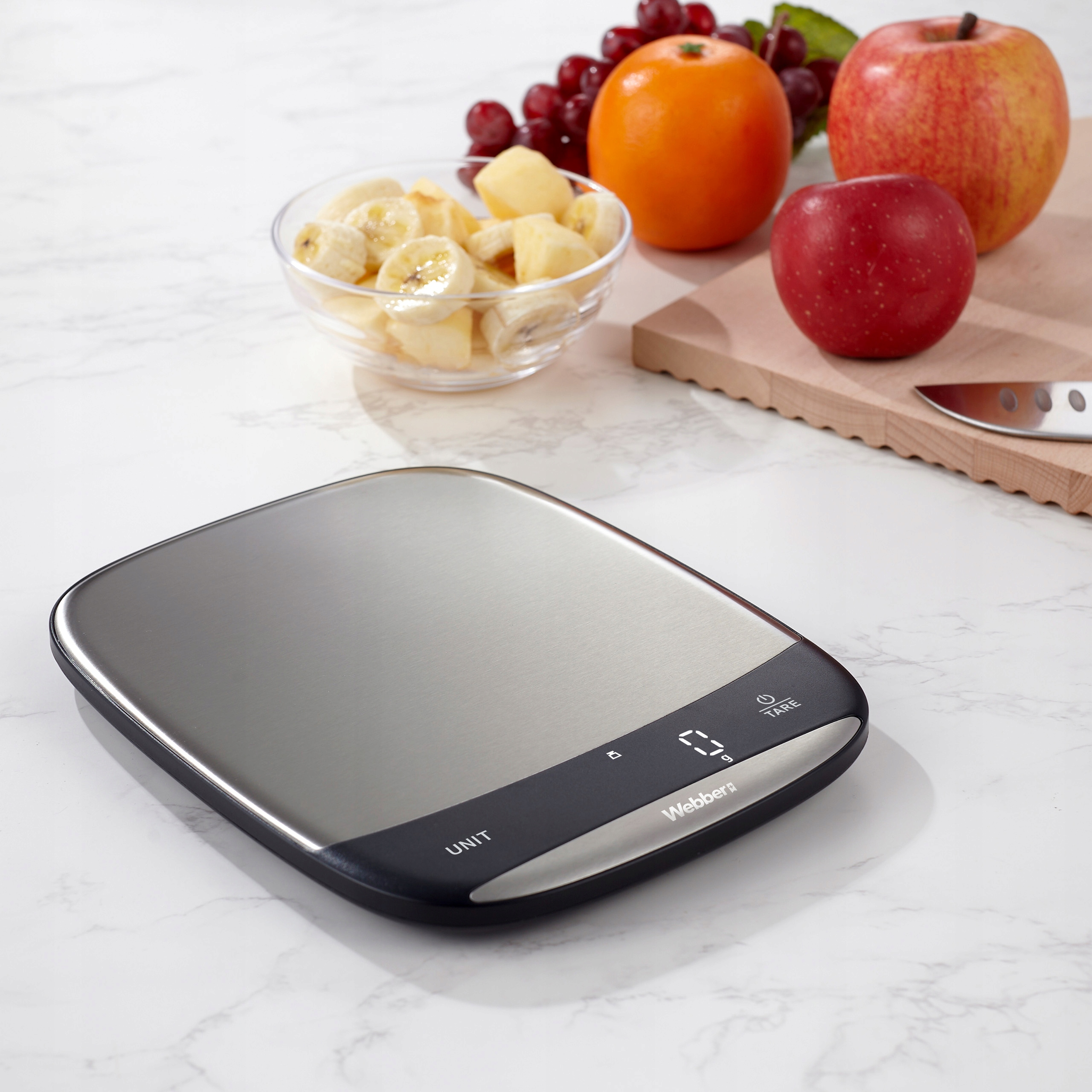 Waga kuchenna z owocami na marmurowej powierzchni. Zawiera banany, winogrona, pomarańcze i jabłka.