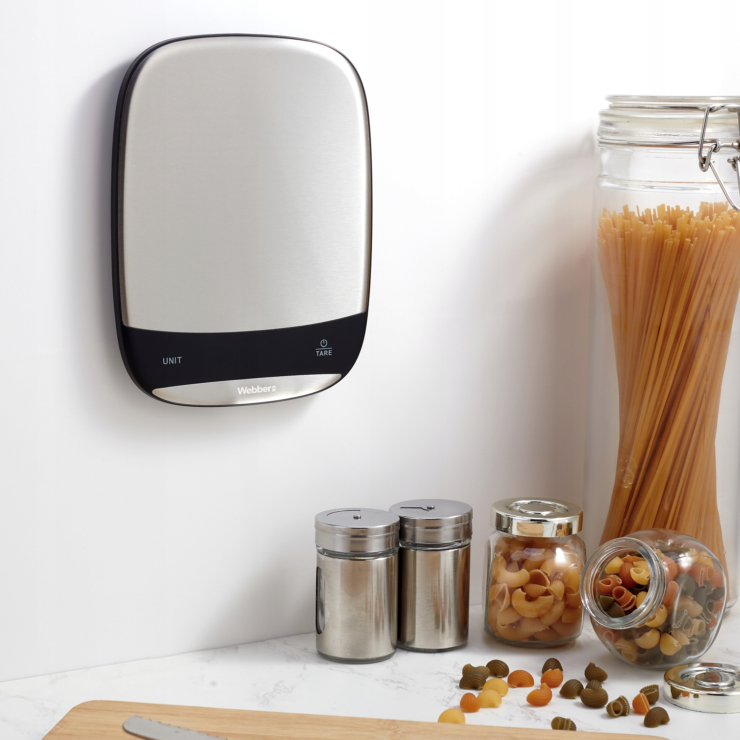 Scena kuchenna z wagą ścienną, słoikami makaronu i przyprawami na marmurowym blacie. Deska do krojenia z nożem.