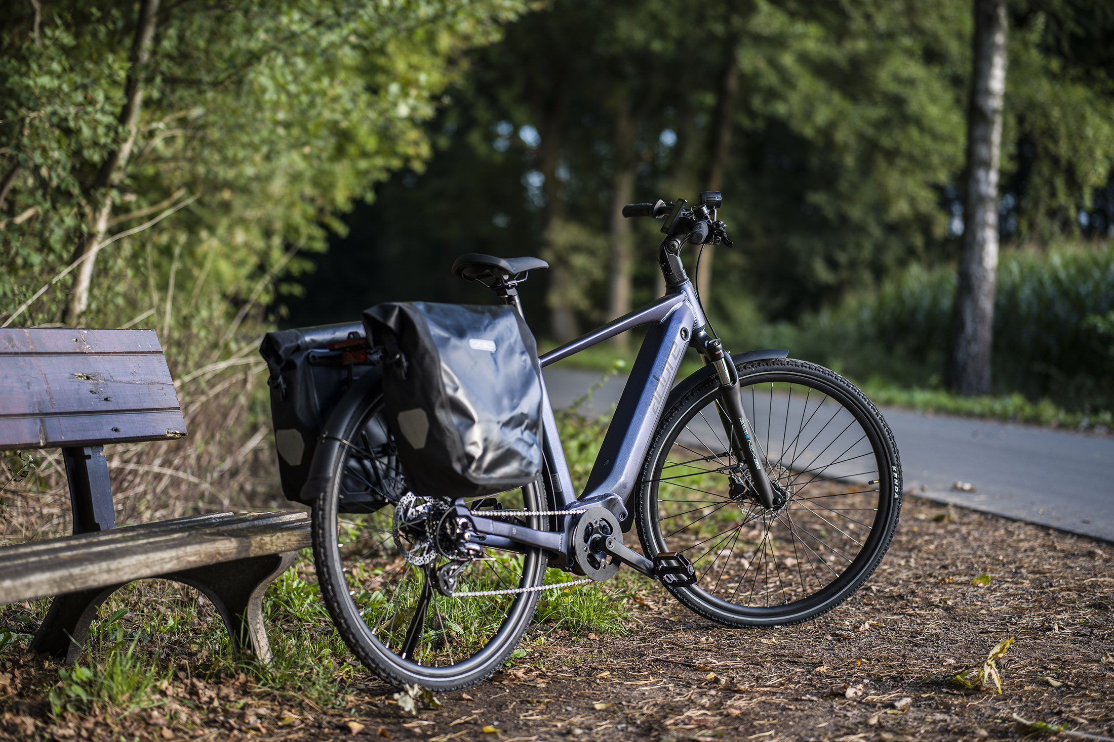 Ein Elektrofahrrad steht in der Nähe einer Holzbank auf einem Weg. Bäume und Büsche umgeben die Szene, eine asphaltierte Straße ist sichtbar.