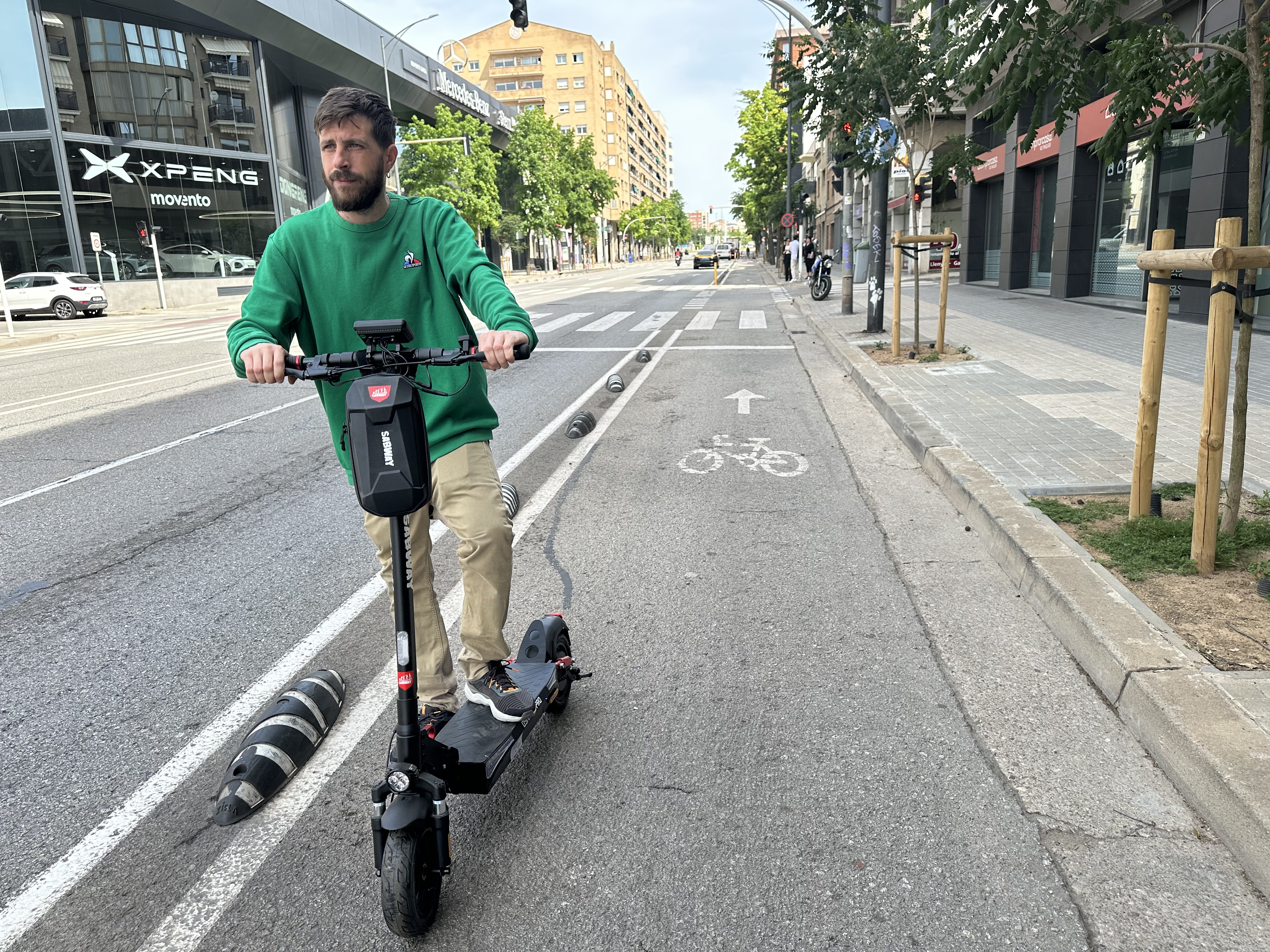 Un hombre de verde monta un patinete eléctrico en una calle. Edificios y árboles bordean la carretera.