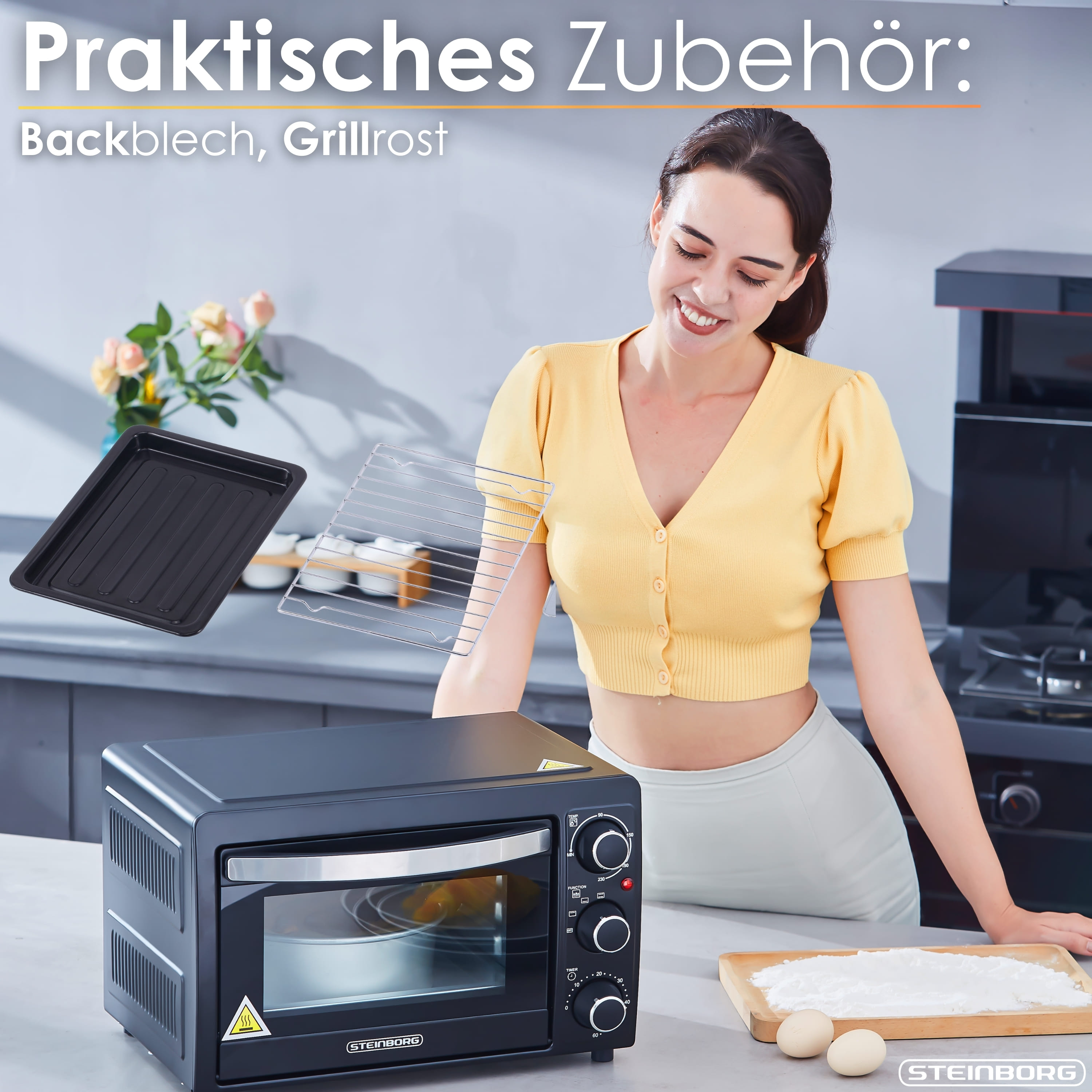 Eine Frau lächelt neben einem schwarzen Ofen mit Zubehör. Ein Backblech und ein Grill sind in der Luft. Eier und Mehl auf einem Brett.