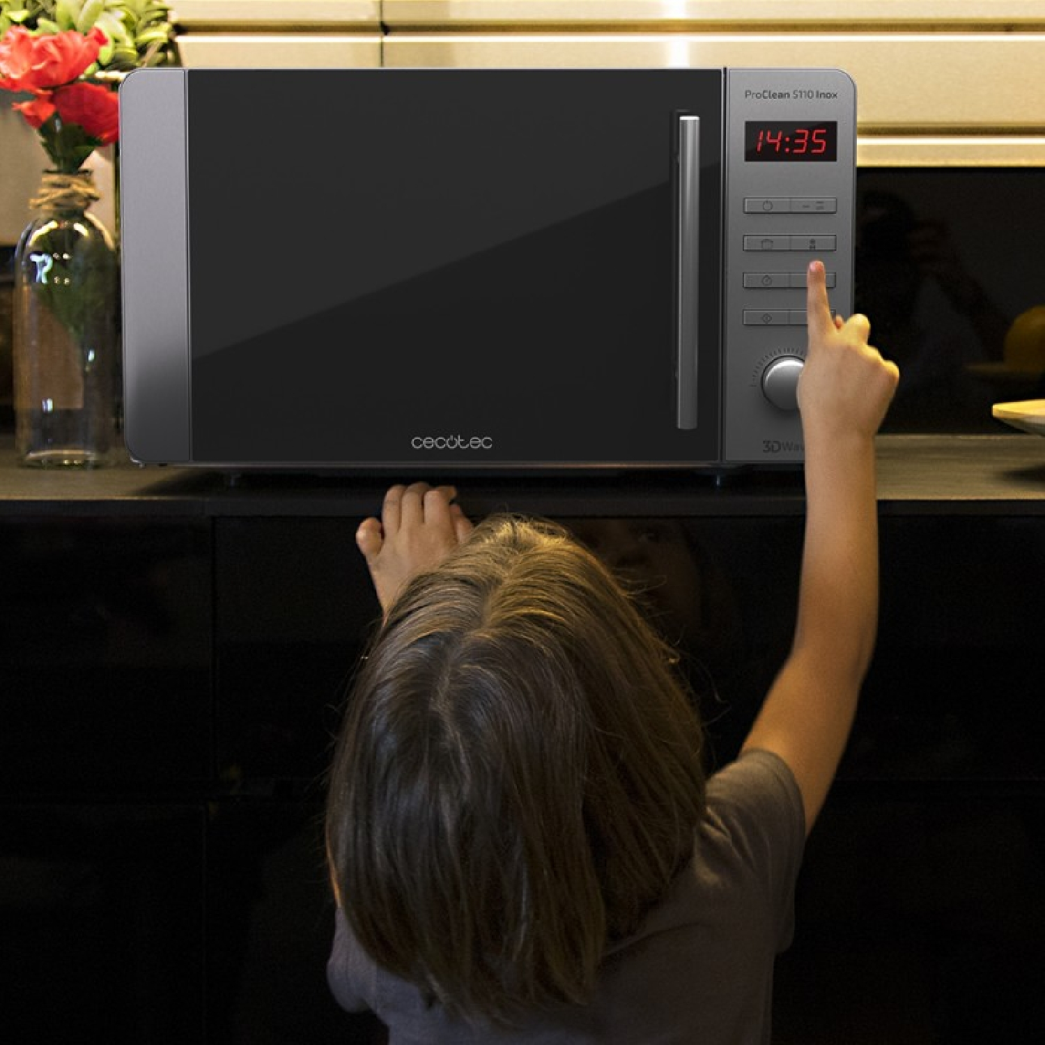 Dziecko sięga, aby nacisnąć przycisk na kuchence mikrofalowej. Kuchenka mikrofalowa stoi na czarnym blacie. Wazon z kwiatami po lewej.