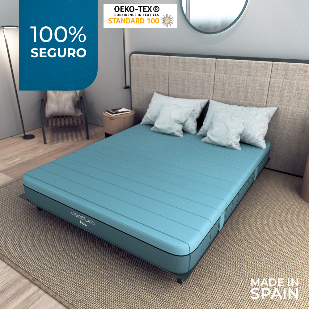 Un colchón azul con almohadas en una cama. La cama está en una habitación con una alfombra y una mesita de noche. Texto dice 'Made in Spain'.