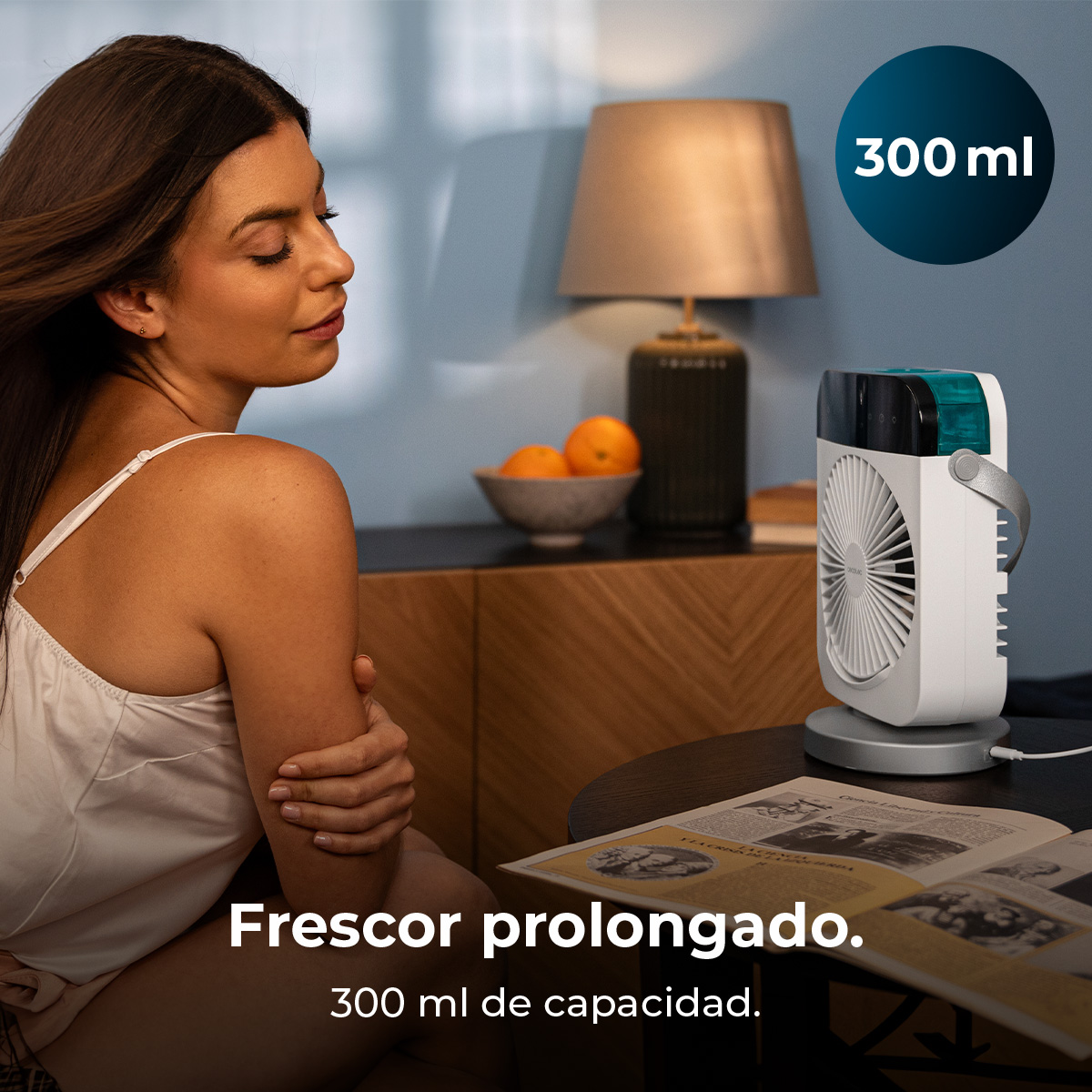 Mujer sentada cerca de un ventilador. Un cuenco de naranjas y una lámpara están sobre una mesa. El ventilador es blanco con la parte superior turquesa.