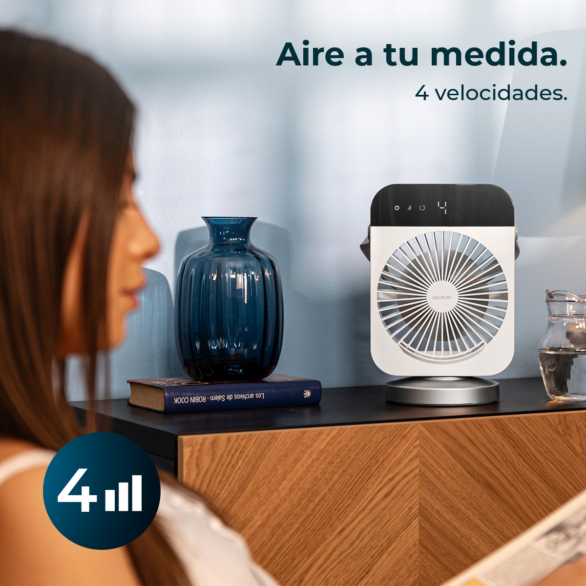Una mujer mira un ventilador sobre una mesa con un jarrón azul y un libro. El texto dice 'Aire a tu medida. 4 velocidades'.