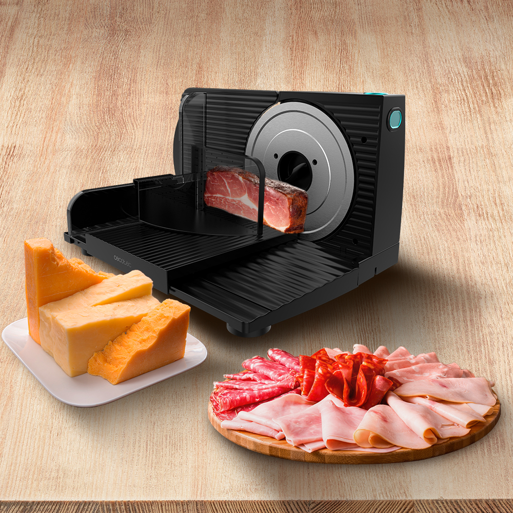 Un'affettatrice con prosciutto, formaggio e salumi su un tagliere di legno. Un blocco di formaggio è su un piatto bianco.