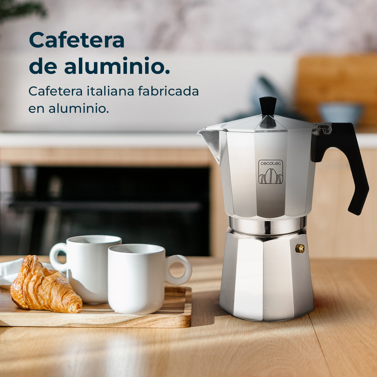 Una cafetera plateada sobre una mesa de madera con dos tazas blancas y un cruasán. El fondo muestra un ambiente de cocina.