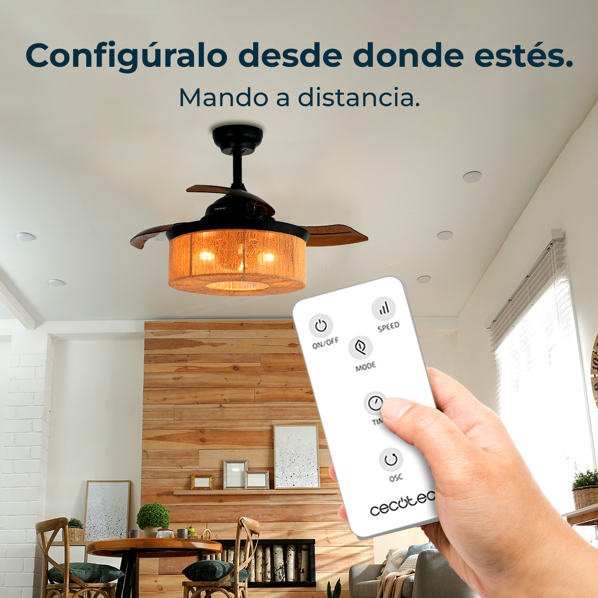 Una mano sostiene un mando a distancia. Un ventilador de techo con luz arriba. Una pared de madera y muebles en el fondo.