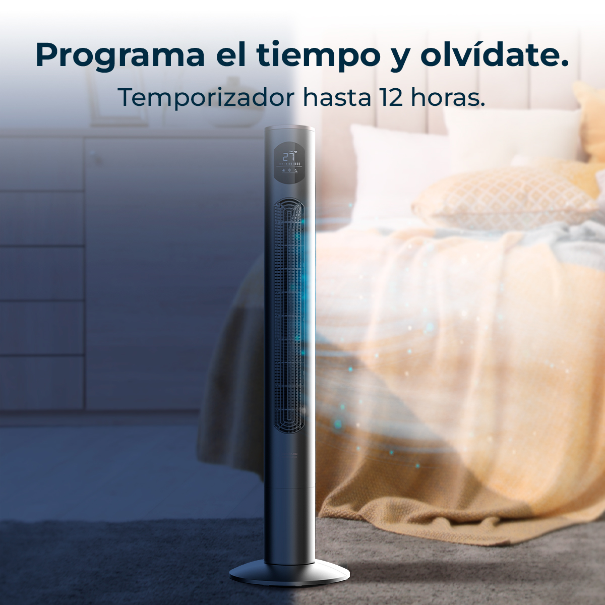 Un ventilador de torre plateado está en un dormitorio. El ventilador muestra 27 grados. Una cama con almohadas y una manta está en el fondo.