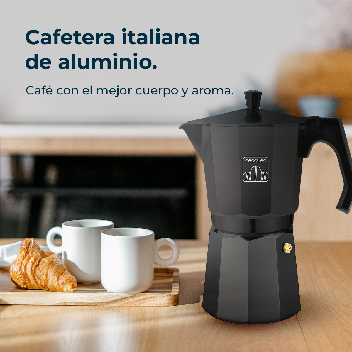 Una cafetera italiana negra sobre mesa de madera. Dos tazas blancas y un cruasán están cerca. Texto en español visible.
