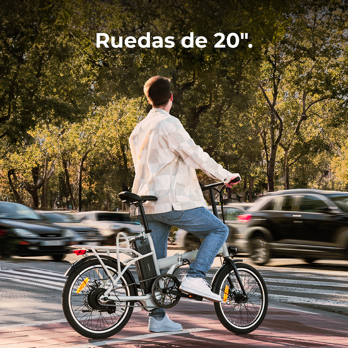 Una persona in bicicletta attraversa le strisce pedonali. Auto sfocate sullo sfondo. Alberi e testo "Ruedas de 20".