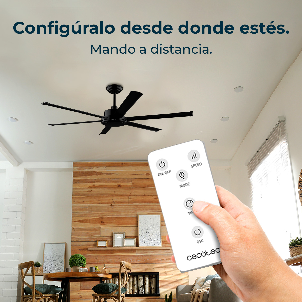 Una mano sostiene un mando a distancia, controlando un ventilador de techo negro. El fondo muestra una sala de estar con pared de madera y muebles.