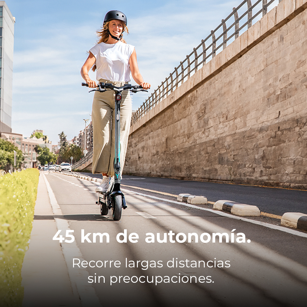 Una mujer monta un patinete eléctrico en una carretera. Lleva casco y sonríe. Un muro de ladrillo y edificios están al fondo.