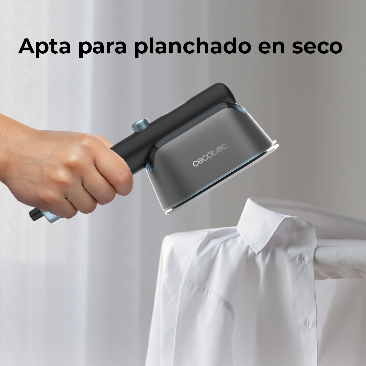 Una mano tiene un ferro da stiro nero e grigio sopra una camicia bianca su un'asse da stiro. Lo sfondo è una tenda bianca.
