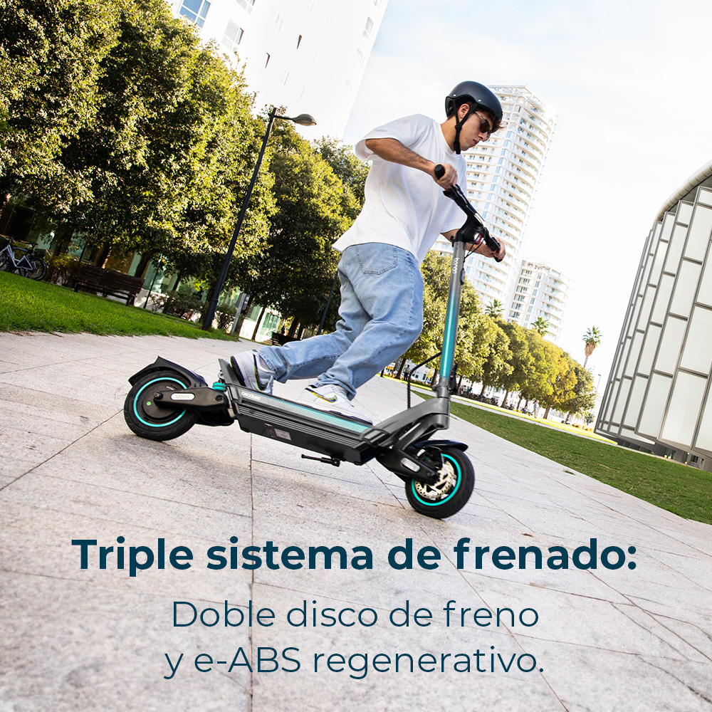 Un hombre monta un patinete eléctrico en una acera. Lleva casco. Edificios y árboles al fondo.