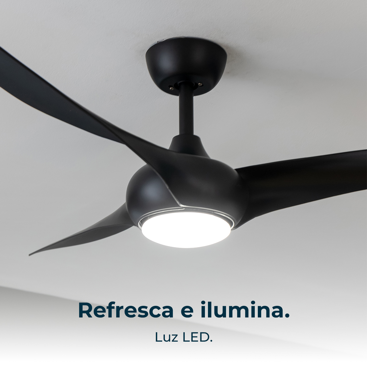 Un ventilatore da soffitto nero con tre pale e una luce LED centrale è montato su un soffitto bianco. Testo: Refresca e ilumina.